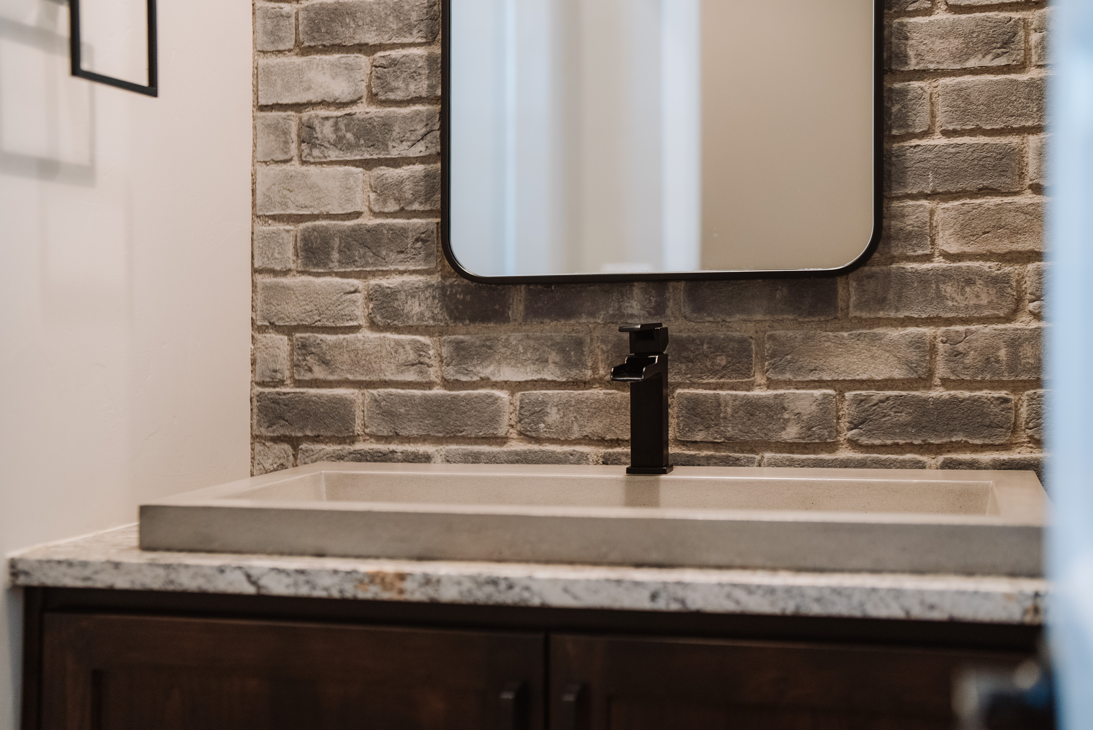 This bathroom features a contemporary design highlighted by a rustic gray brick accent wall and a sleek, rectangular concrete vessel sink. A modern black faucet adds a sophisticated touch, perfectly coordinating with the minimalist black-framed mirror above. The dark wood cabinetry and speckled granite countertop create a refined, industrial-chic aesthetic.