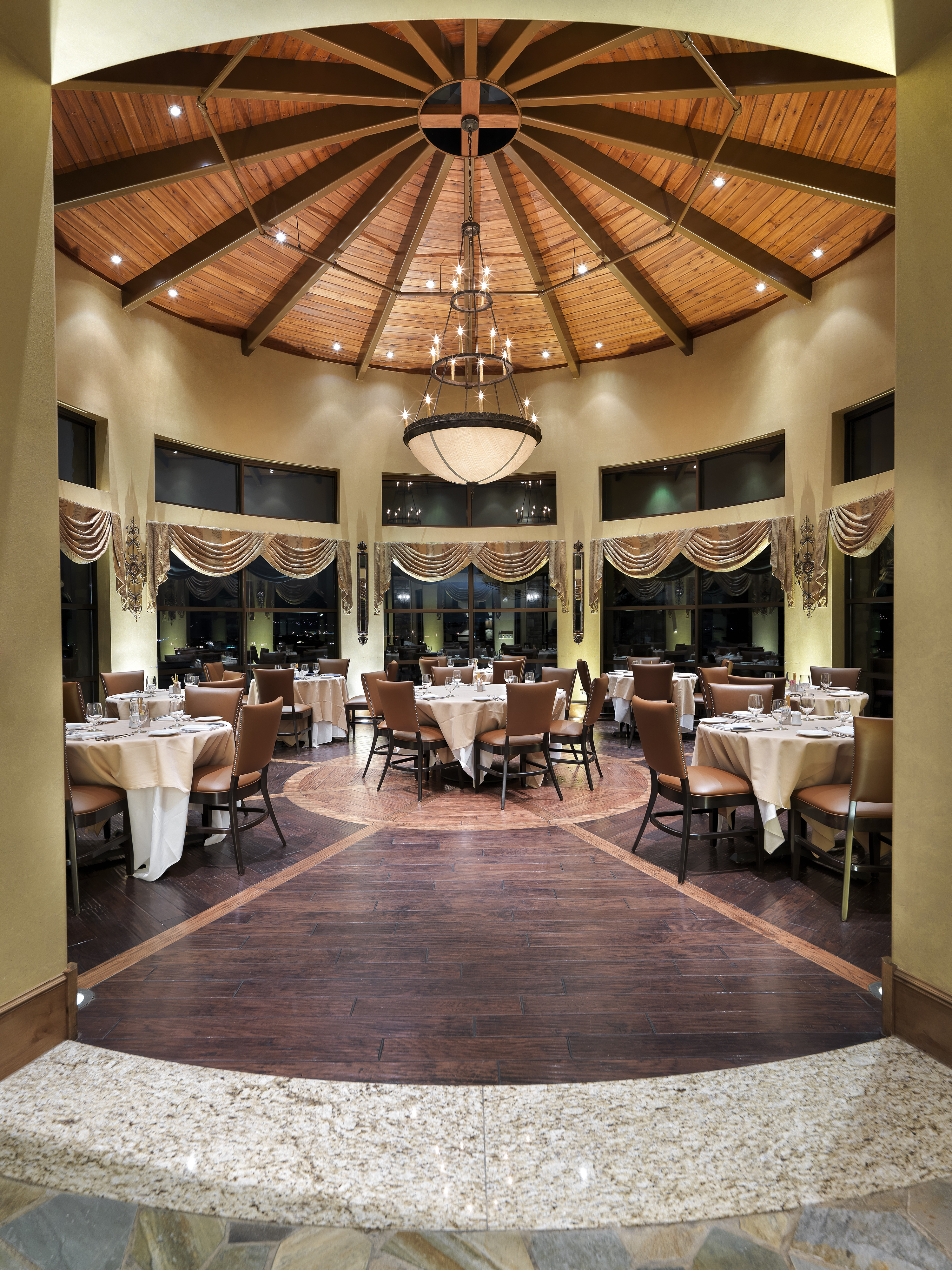This elegantly appointed circular dining room features a stunning vaulted wood-paneled ceiling with a central chandelier, creating a grand and inviting atmosphere. Multiple tables with tan tablecloths and leather chairs are arranged elegantly beneath large windows draped in formal valances. The rich, dark hardwood floors and sophisticated architectural geometry make this space perfect for upscale event hosting or fine dining.