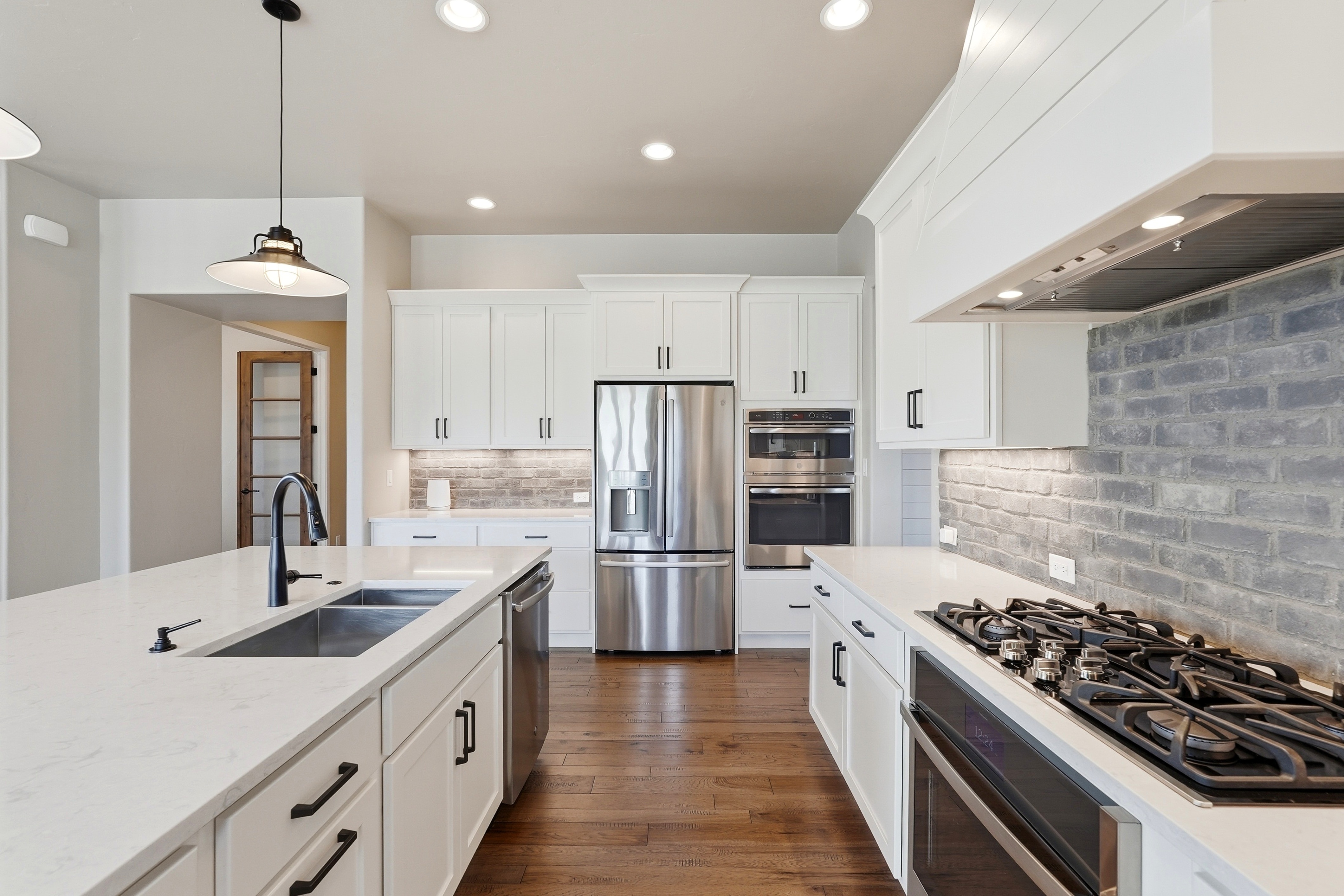 This modern kitchen features crisp white cabinetry, high-end stainless steel appliances, and elegant quartz countertops throughout. A large central island provides ample workspace, while a distinctive gray brick backsplash adds texture and character against the clean aesthetic. The warm wood flooring and tasteful pendant lighting create a welcoming and well-appointed space perfect for entertaining.