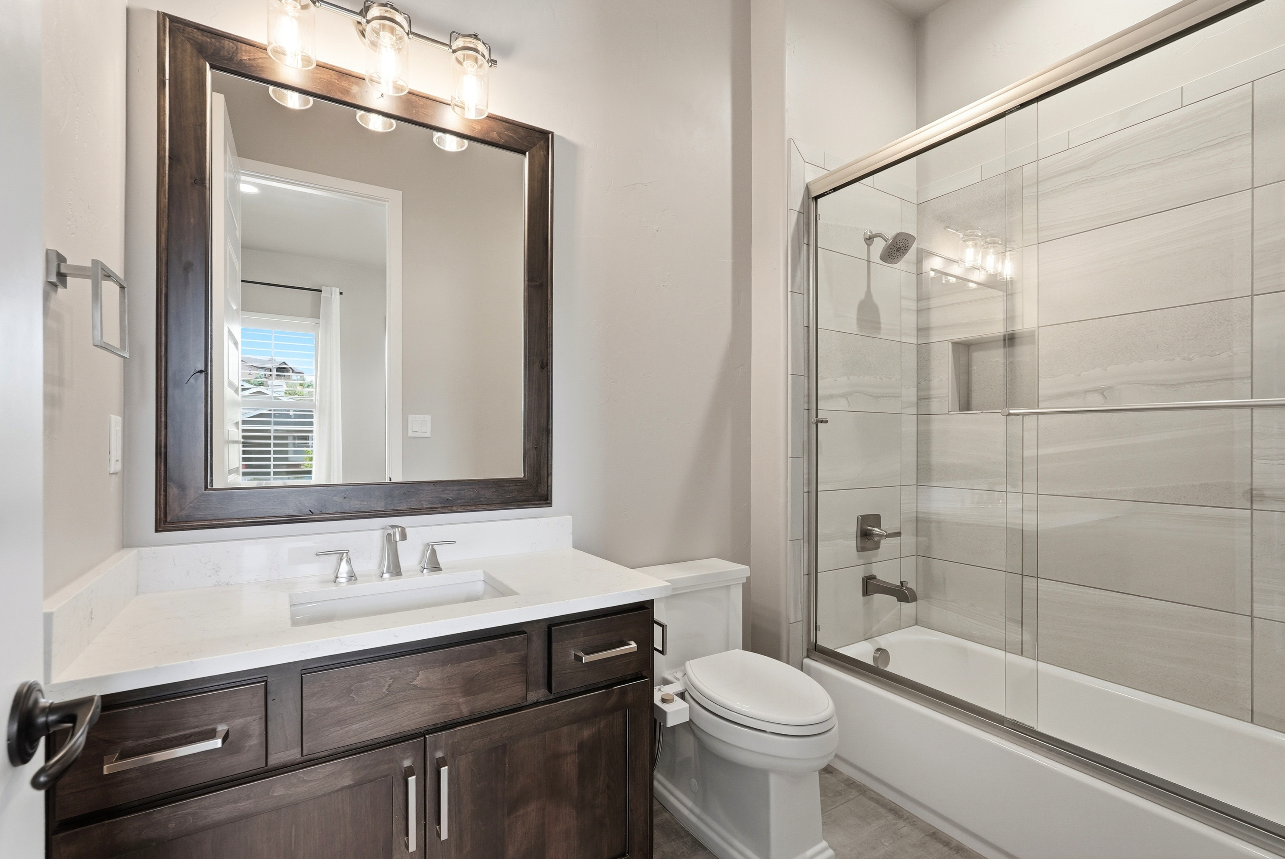 This modern bathroom features a dark wood vanity with a white quartz countertop and a contemporary undermount sink. The space is equipped with a combination shower and bathtub enclosure with sleek tile surround, glass doors, and polished chrome fixtures. A large, dark-framed mirror sits above the vanity, complemented by a three-light vanity fixture to create a clean, functional design perfect for a guest space.