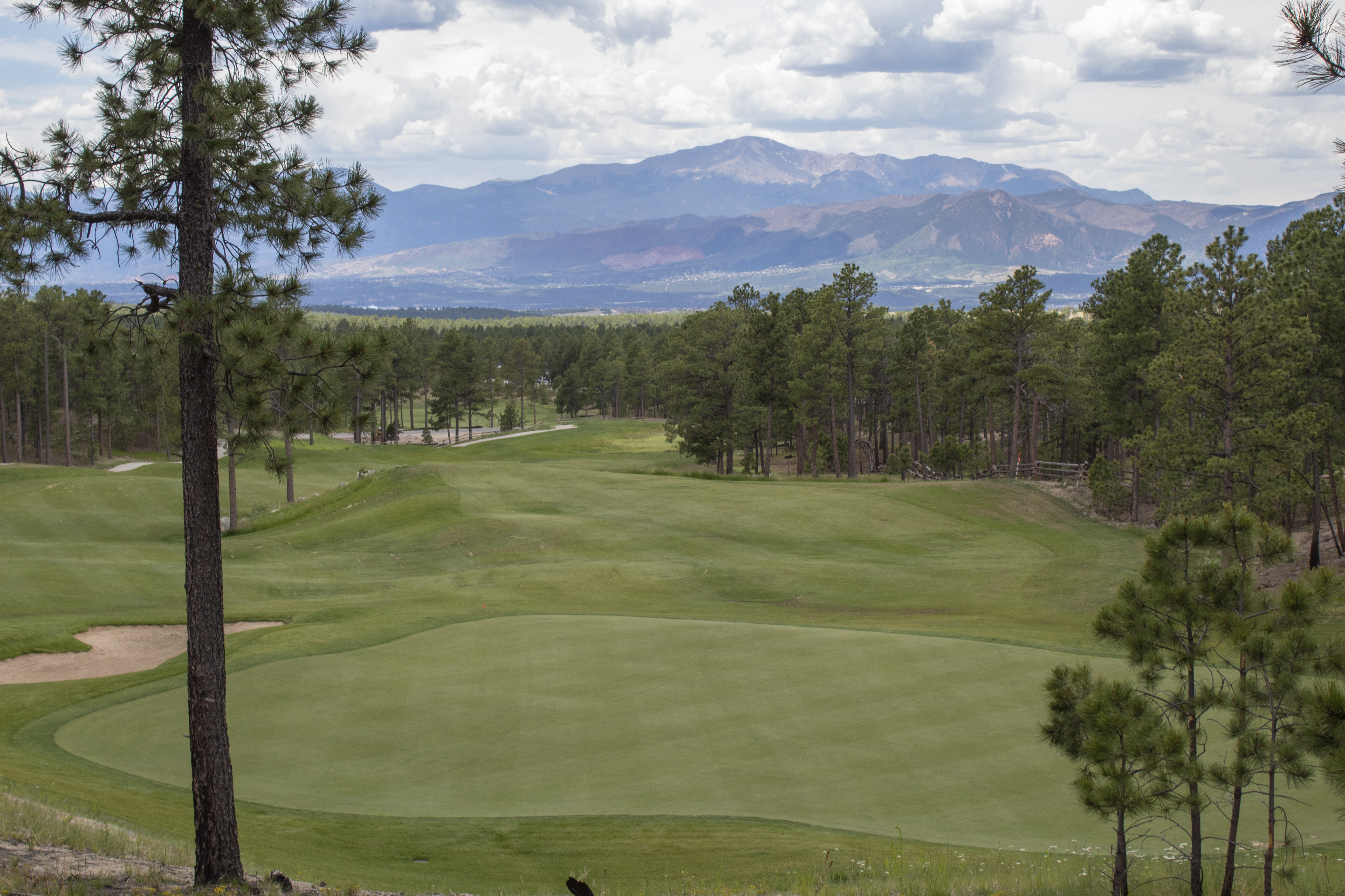 This image showcases a scenic golf course hole surrounded by lush evergreen trees and rolling green hills. A large putting green is prominently featured in the foreground, with the distant mountain range providing a picturesque and tranquil backdrop. This high-end amenity adds significant value, offering residents or club members access to a well-maintained, professionally landscaped recreational space in a beautiful natural setting.
