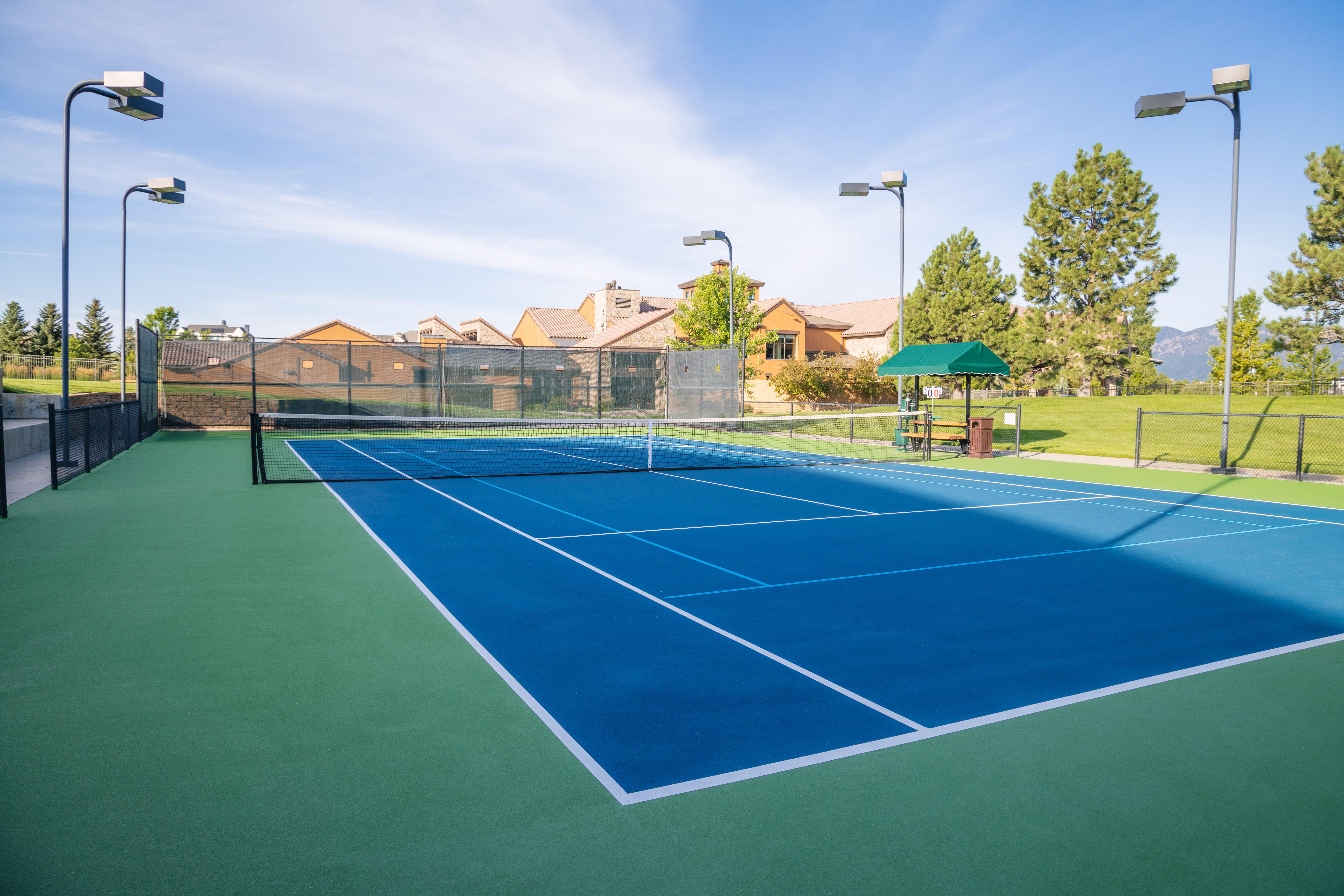 This image features a professionally maintained tennis court with a vibrant blue playing surface and green surrounding areas. The facility includes high-quality lighting, perimeter chain-link fencing, and a shaded seating area, making it a desirable recreational amenity. The setting is clean and well-kept, reflecting a well-managed residential or community property.