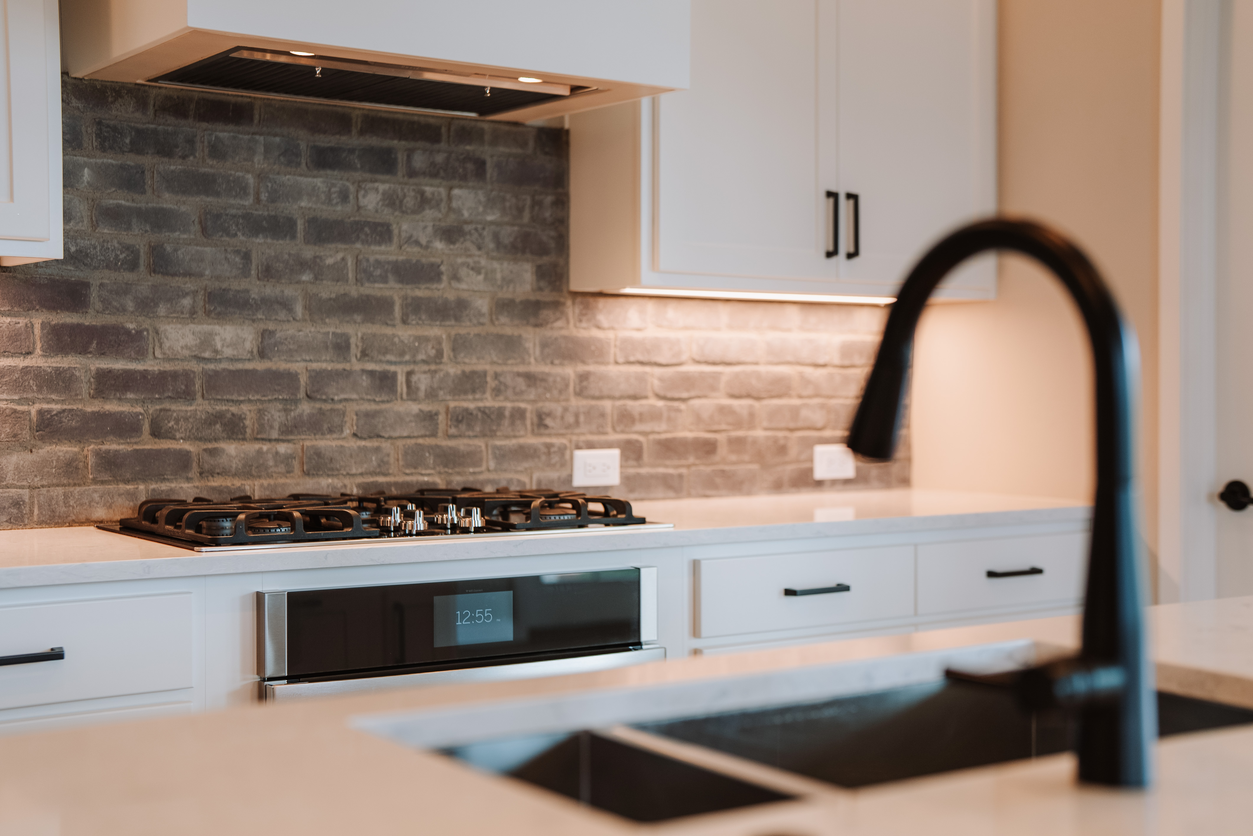 This kitchen features a clean, contemporary design with crisp white cabinetry and a sophisticated grey brick backsplash. A high-end gas cooktop and matching built-in oven are complemented by sleek black hardware and a modern dark faucet. The space is well-lit with under-cabinet lighting, creating a bright and stylish atmosphere perfect for culinary activities.