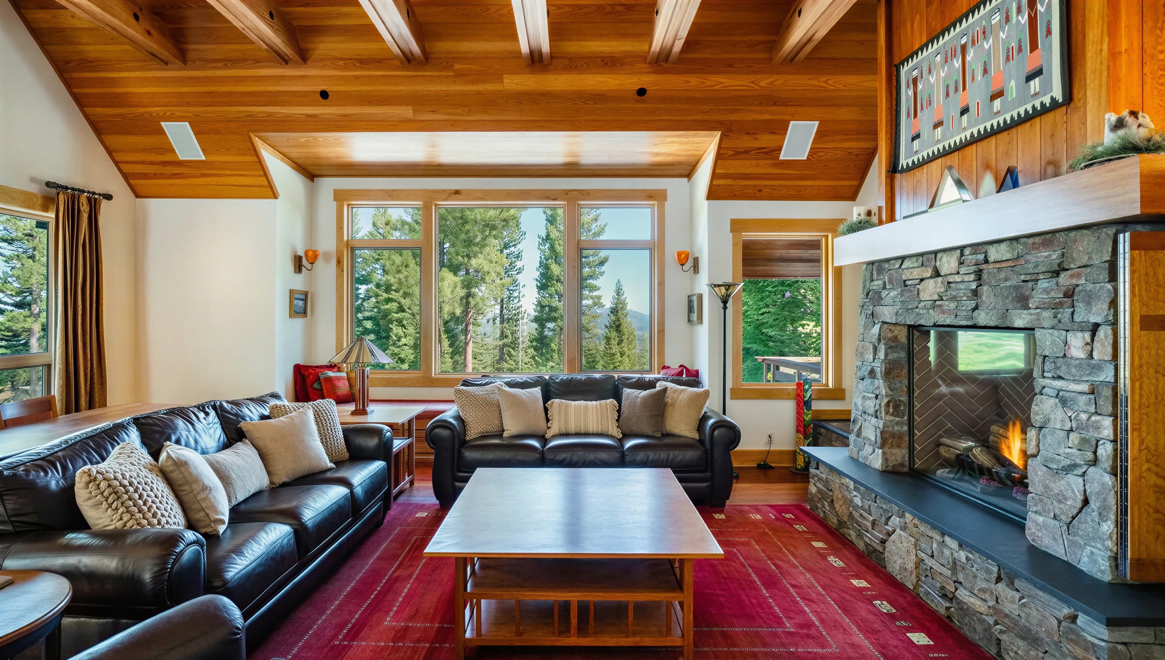 This inviting living room features dual leather sofas facing each other, arranged around a wooden coffee table on a red patterned rug. The focal point is a stone fireplace framed by wooden accents. Natural light floods the room through large picture windows highlighting the vaulted wood beam ceiling, creating a warm and rustic ambiance.