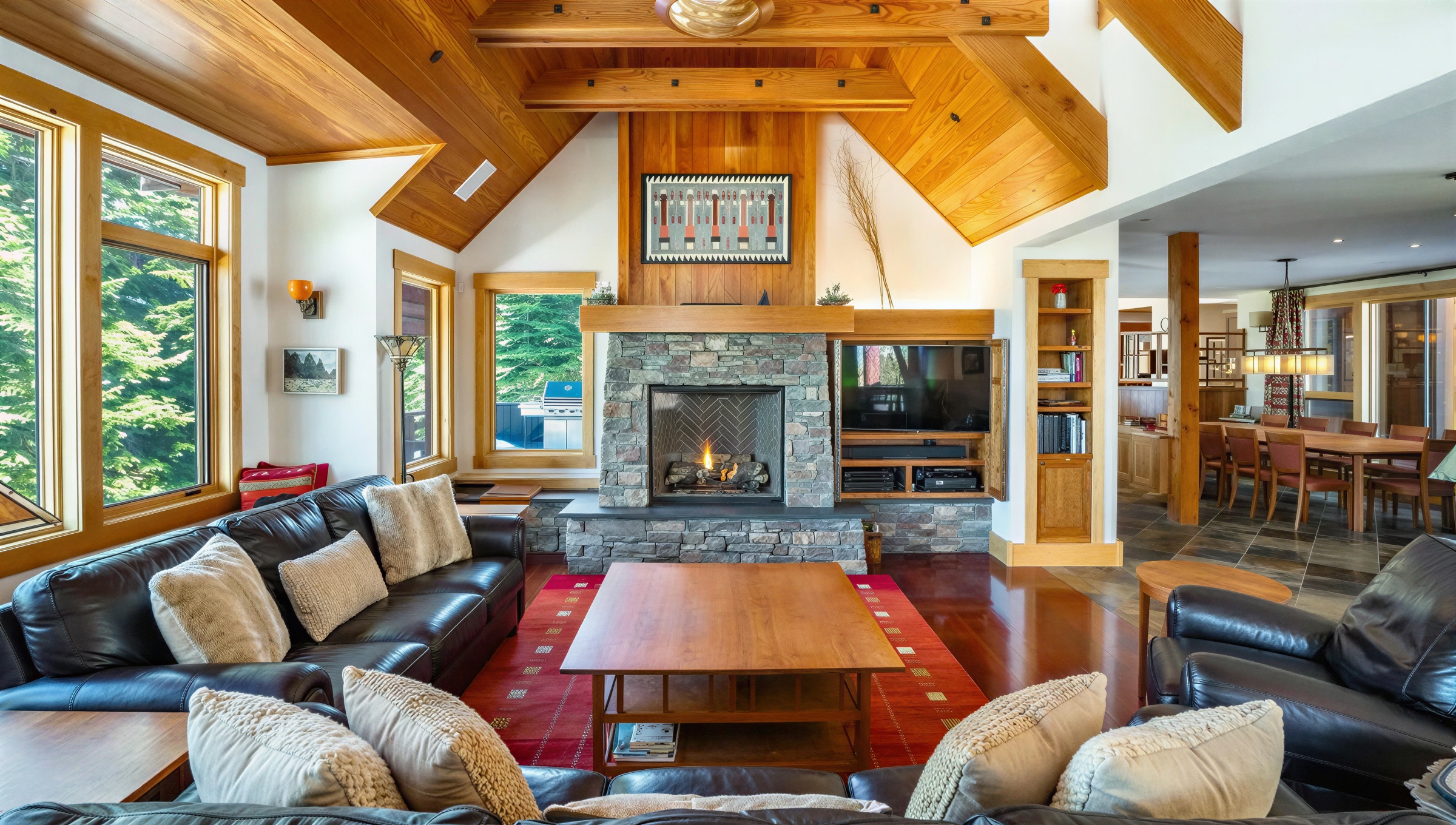 This inviting living room features a stone fireplace with a mounted television, creating a cozy focal point. The room is furnished with comfortable black leather sofas adorned with decorative pillows, centered around a wooden coffee table on a reddish rug. The warm wood finishes on the ceiling and beams add to the home's rustic charm.