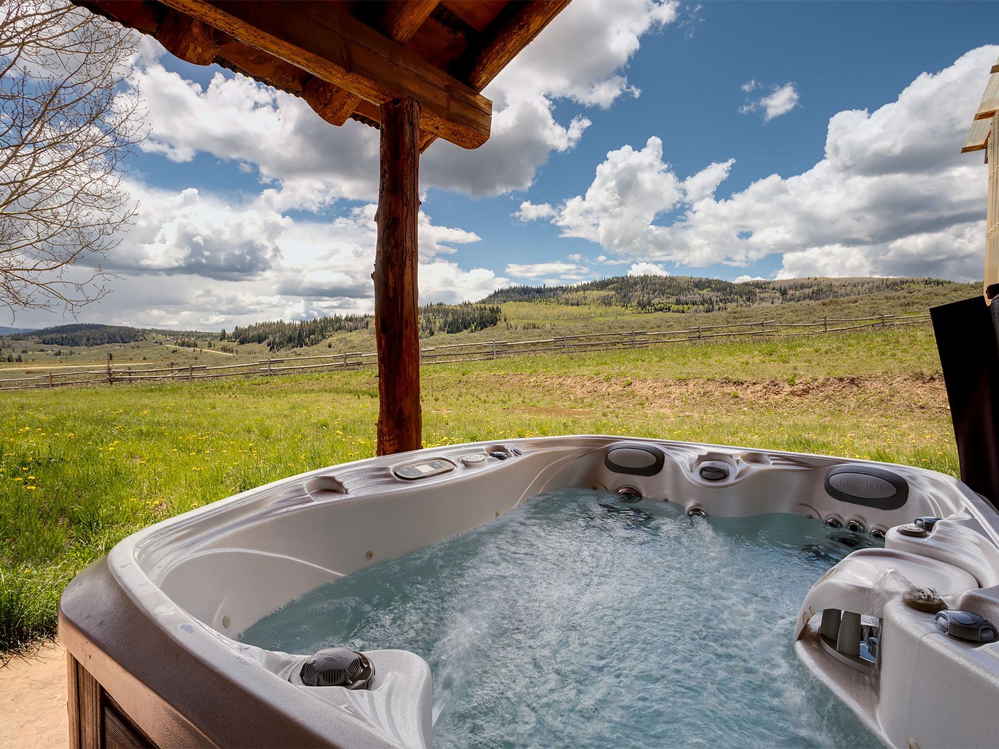 This image showcases a luxurious hot tub with jets operating, situated outdoors in a scenic setting. The hot tub is the central feature, with a wooden porch roof visible overhead. The backdrop includes a grassy field, a distant fence, and tree-covered hills under a partially cloudy sky, creating a relaxing and private atmosphere, ideal for a leisure property.