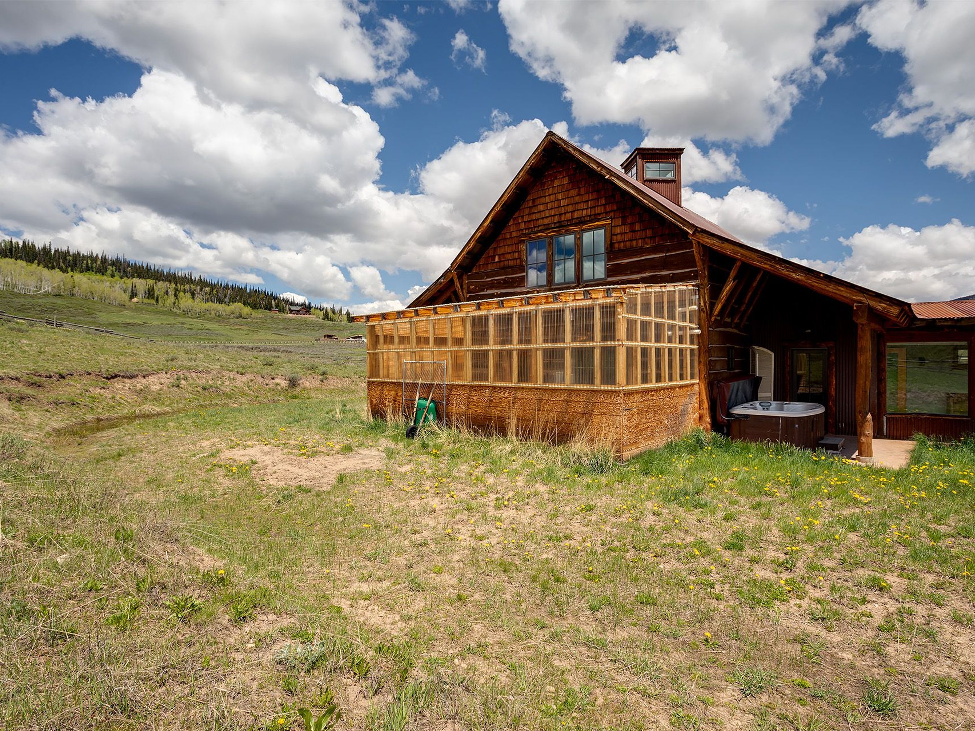 This image showcases the side view of a charming wooden cabin set against a backdrop of a partly cloudy sky. The cabin features a sunroom addition and a hot tub, highlighting the potential for relaxation and enjoyment of the surrounding natural landscape. The grassy yard with scattered yellow flowers adds a touch of vibrancy to the scene.