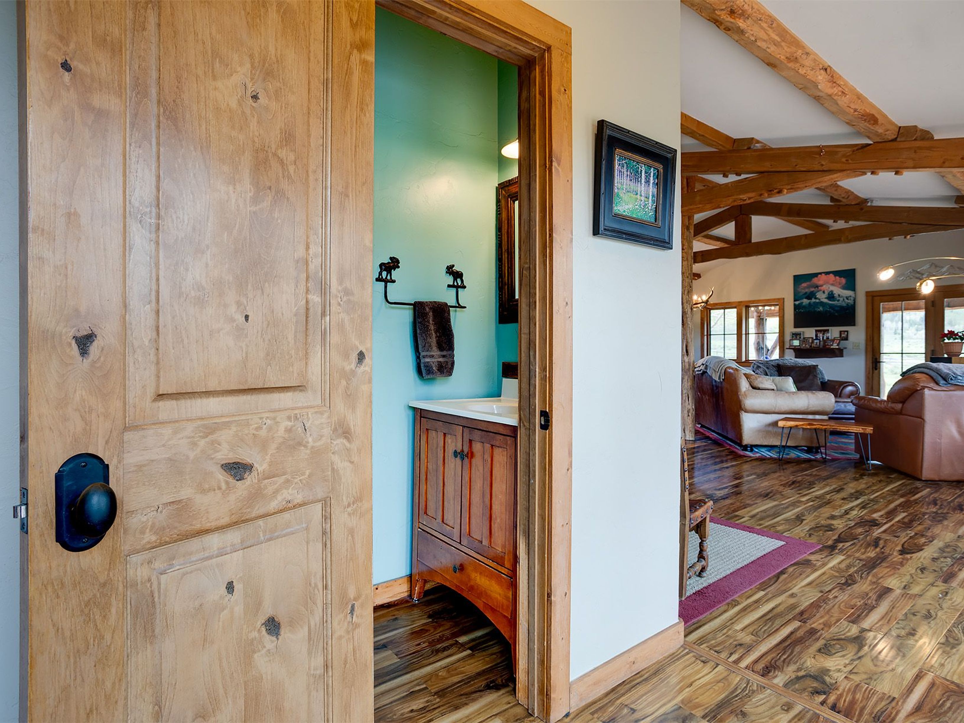 The image shows a bathroom as seen through an open doorway. The bathroom has a wooden vanity with a sink and a towel rack with a towel. The walls are painted a light blue or green color. There is a painting on the wall outside the bathroom, and a living room is visible through another doorway.