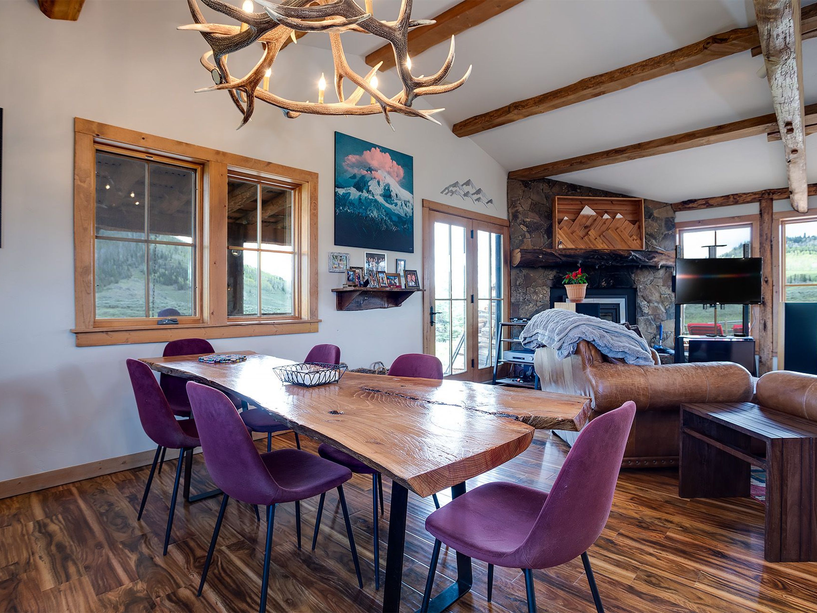 The image showcases a dining room featuring a unique wooden table with natural edges, surrounded by modern purple chairs. An antler chandelier adds a rustic touch, and the room is accented with a mountain-themed artwork and decorative elements. The space transitions into a living area, maintaining a cohesive and inviting atmosphere, suitable for families or entertaining guests.