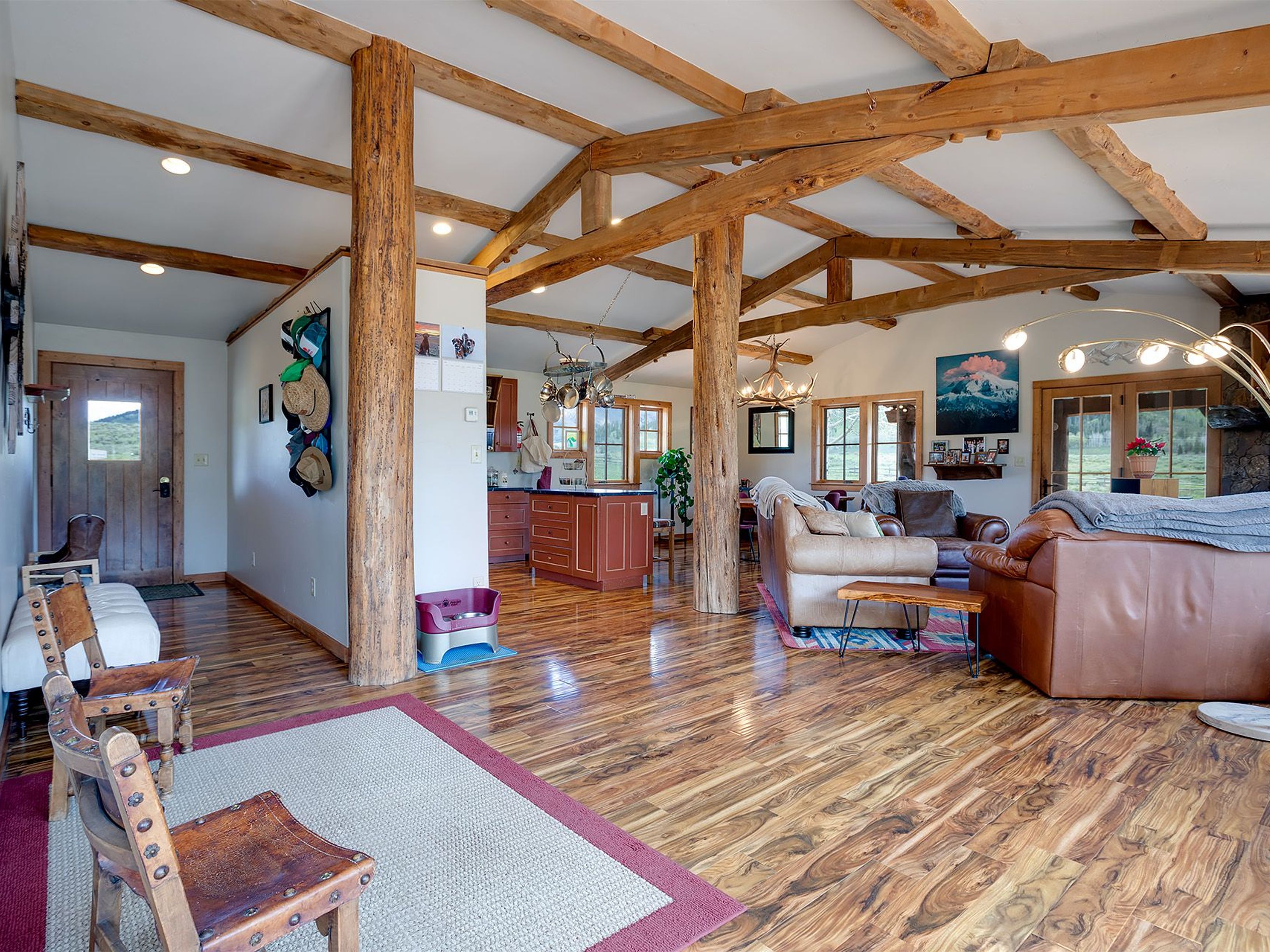 This living room displays a rustic design featuring exposed wooden beams and posts that accent the open floor plan. The hardwood flooring contributes warmth, while comfortable leather sofas provide ample seating. The space connects seamlessly to the kitchen area, creating an inviting atmosphere for relaxation and entertaining.