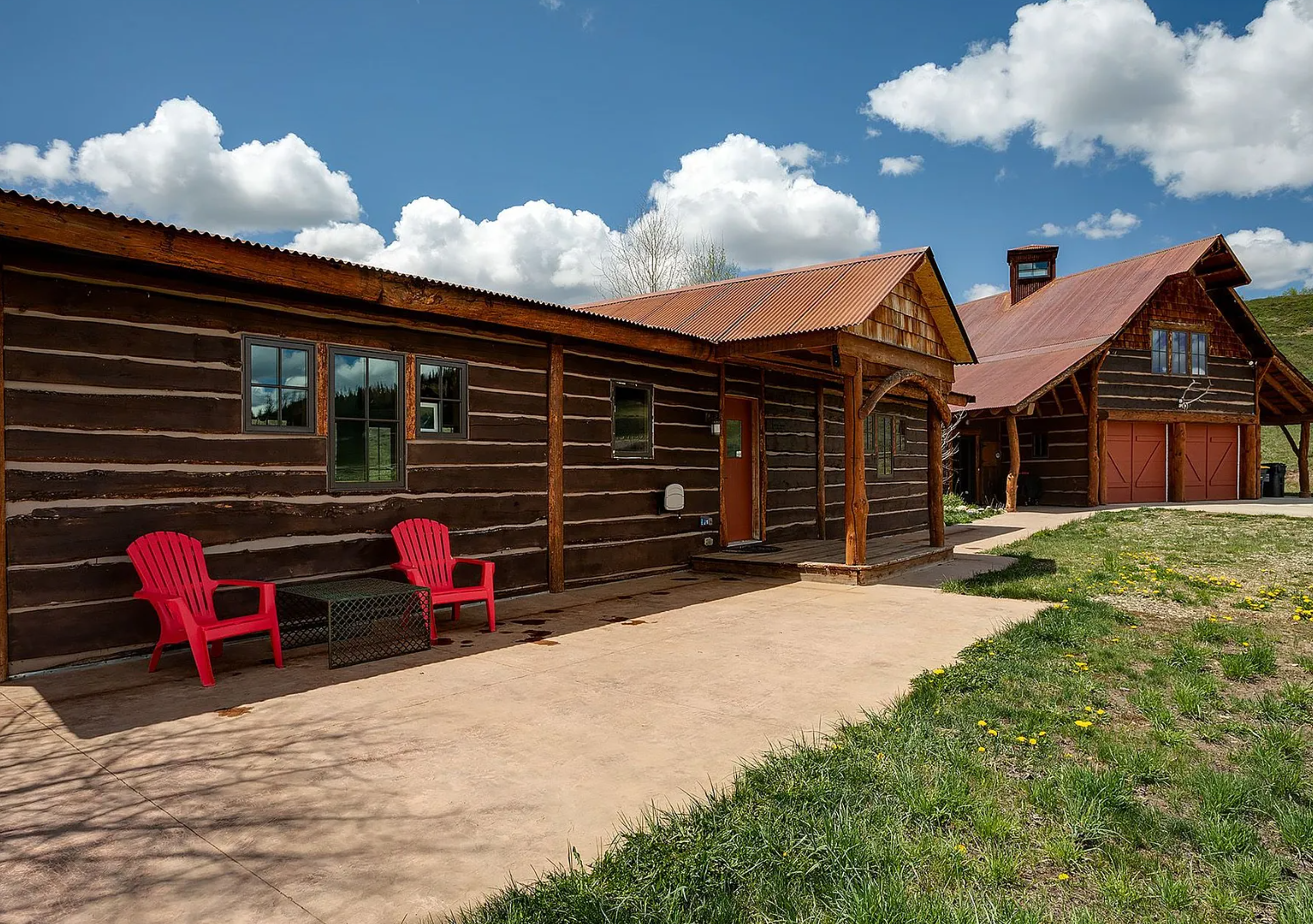 The image showcases the front exterior of a charming log cabin-style home. The structure features a rustic facade with a corrugated metal roof, complemented by a concrete patio and two red chairs providing inviting curb appeal. A secondary, larger cabin structure with garage doors can be seen in the background creating the impression of a spacious property.