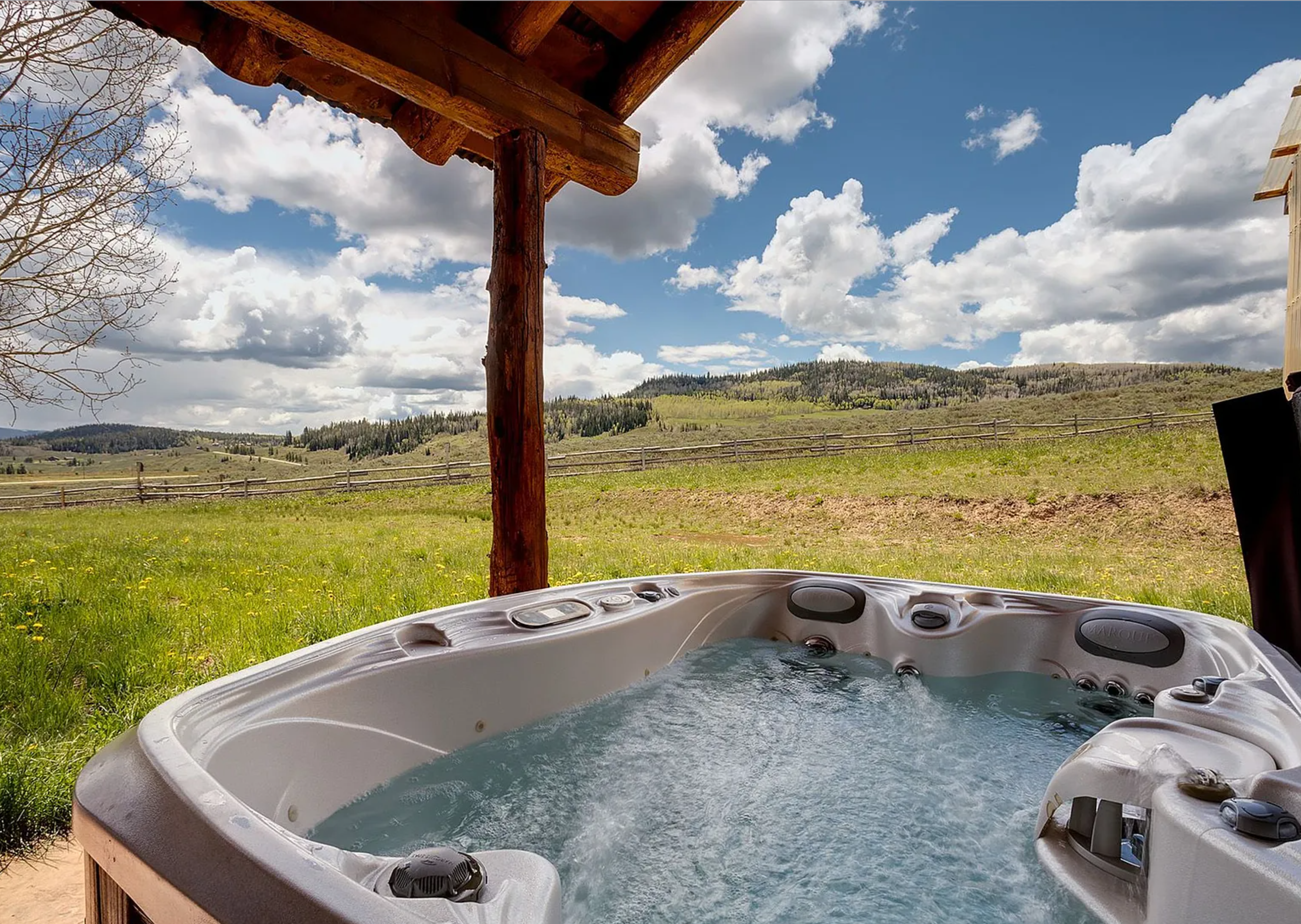 This image showcases a luxurious hot tub set against a serene landscape. The tub features a light-colored shell and crystal-clear water, complemented by a picturesque backdrop of rolling hills and a partly cloudy sky. A rustic wooden structure provides partial coverage, enhancing the spa-like atmosphere.