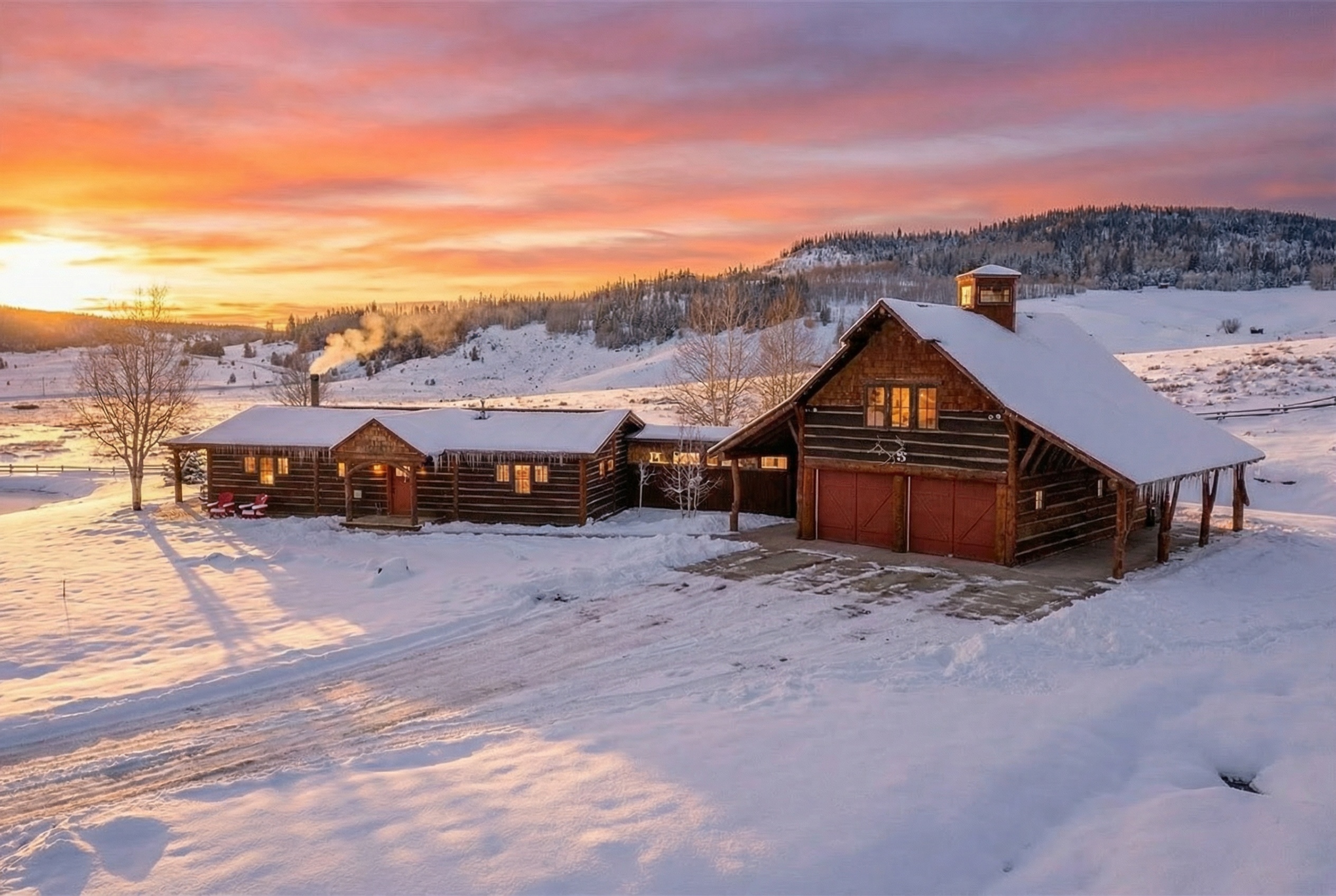 This is a picturesque front view of a log cabin style home and a barn, covered in snow, set against a vibrant sunset. The main house features multiple connected sections with a charming rustic aesthetic, including a smoking chimney. The barn has large red garage doors, and the entire property is blanketed in a pristine layer of snow, creating a serene and inviting atmosphere suitable for a winter retreat or a mountain property listing.