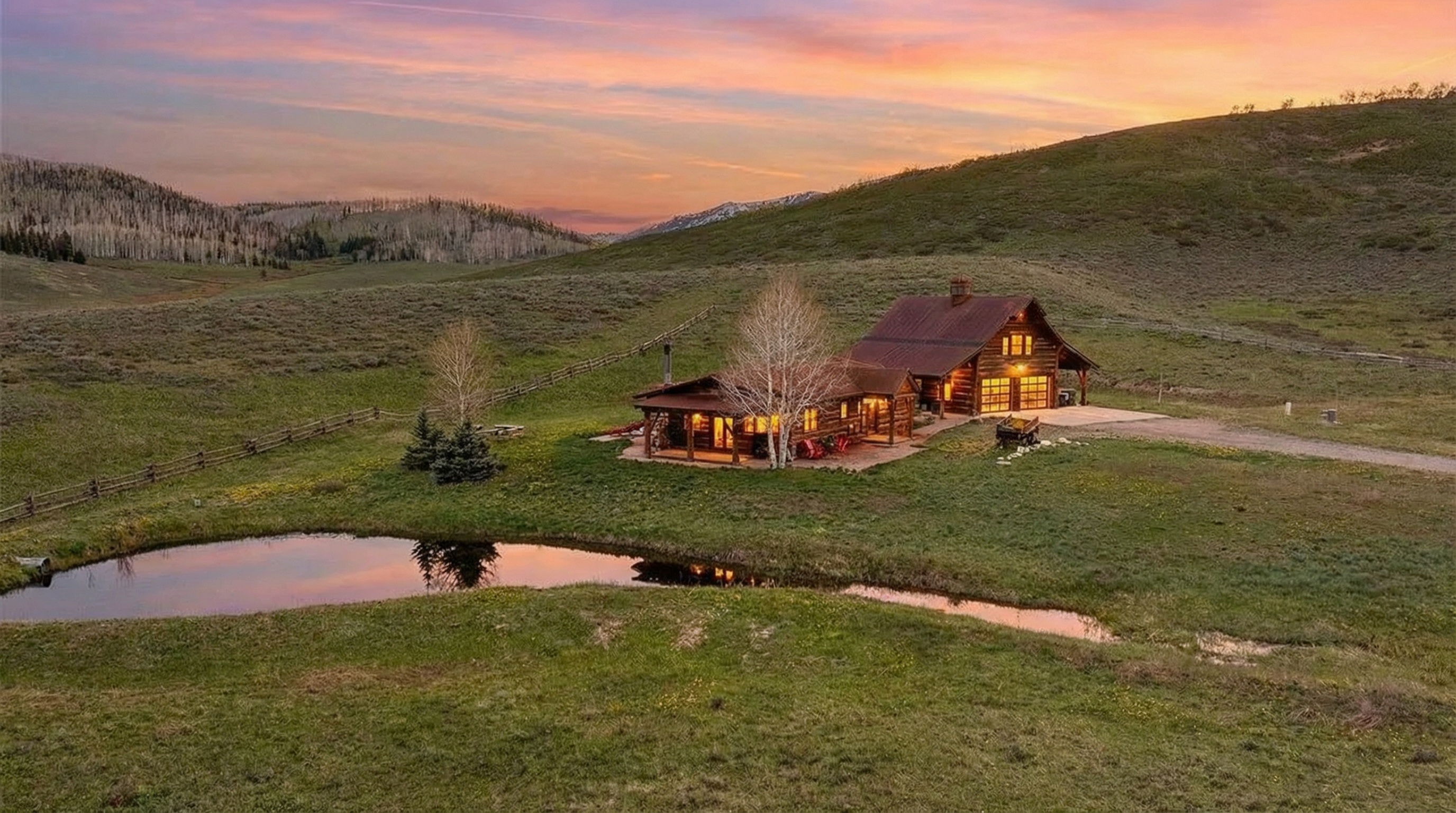 This aerial shot showcases a beautiful log cabin home nestled in a serene landscape. The property features a tranquil pond reflecting the evening sky, lush green fields, and gentle rolling hills in the background. The home's warm interior lighting adds to the inviting atmosphere, making it an ideal retreat.