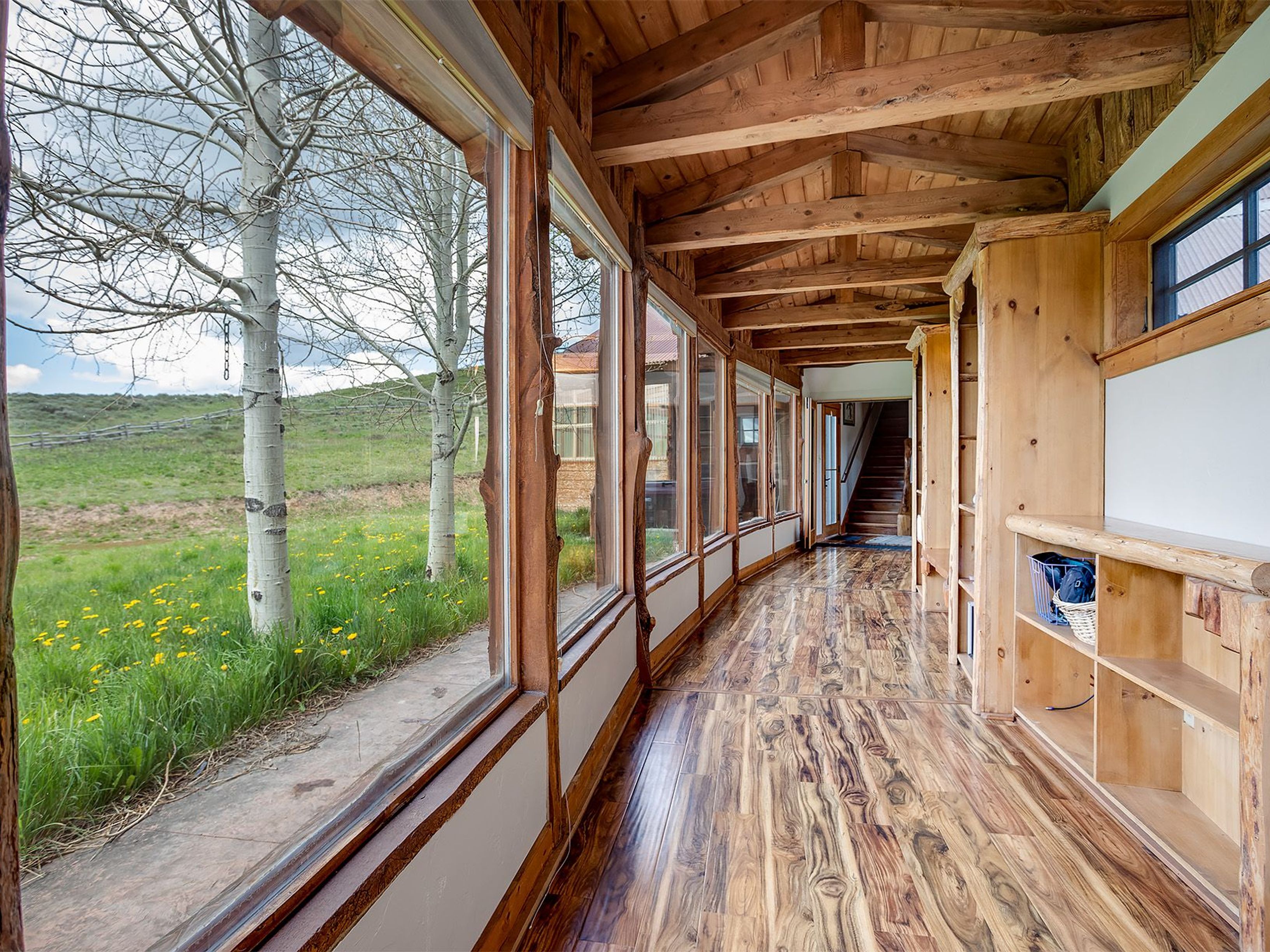 The image showcases a unique hallway/stair area featuring extensive wood finishes. Large picture windows reveal an outside view of a grassy field with aspen trees. The hardwood flooring and exposed wood beams on the ceiling add a rustic charm to the space.