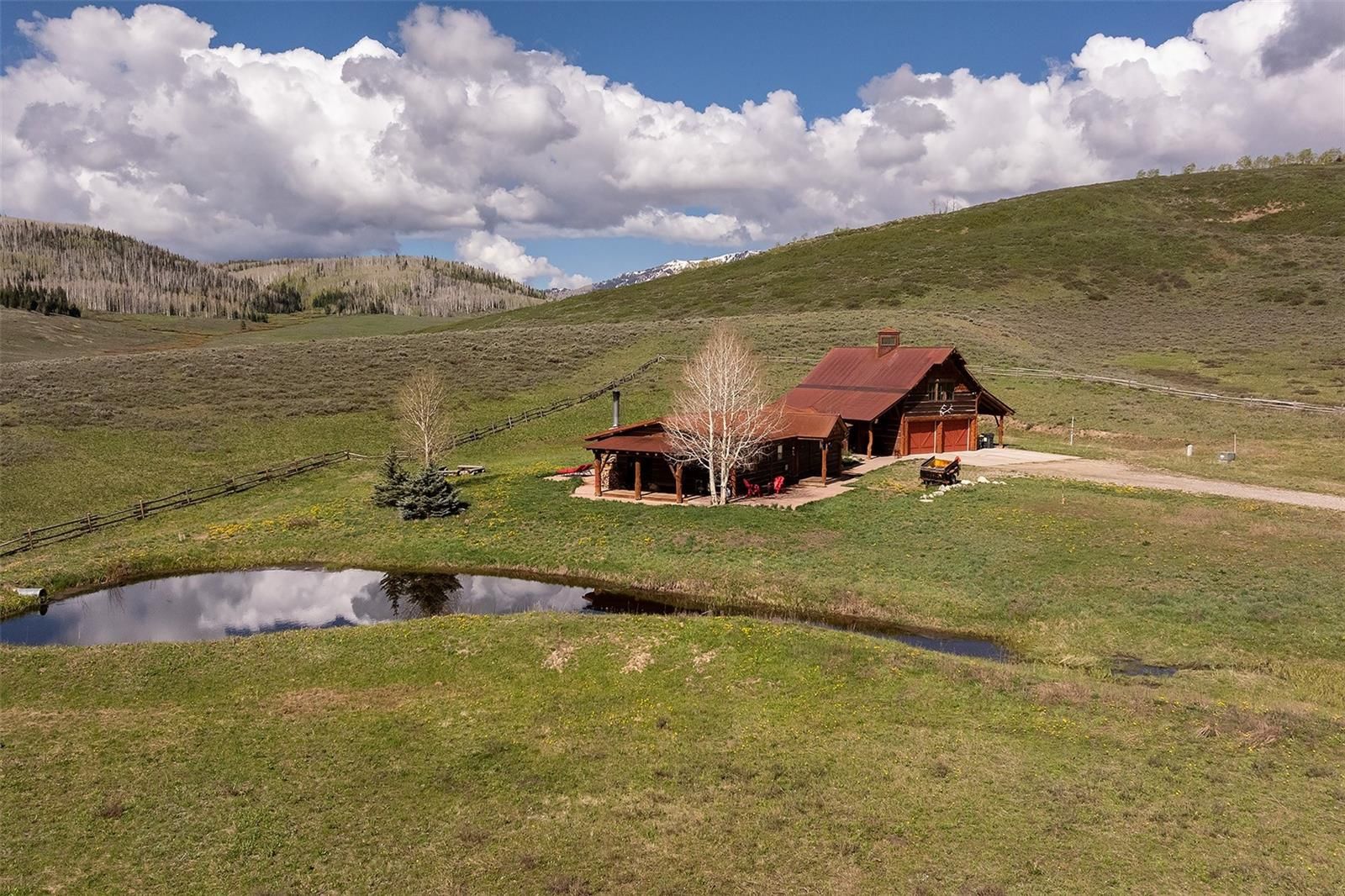 This aerial shot presents a rustic home with a red metal roof, nestled in a rolling green landscape. The property features a small pond in the foreground, a detached structure with a porch, a gravel driveway, and a backdrop of hills and scattered trees. The overall impression is one of secluded, pastoral charm.