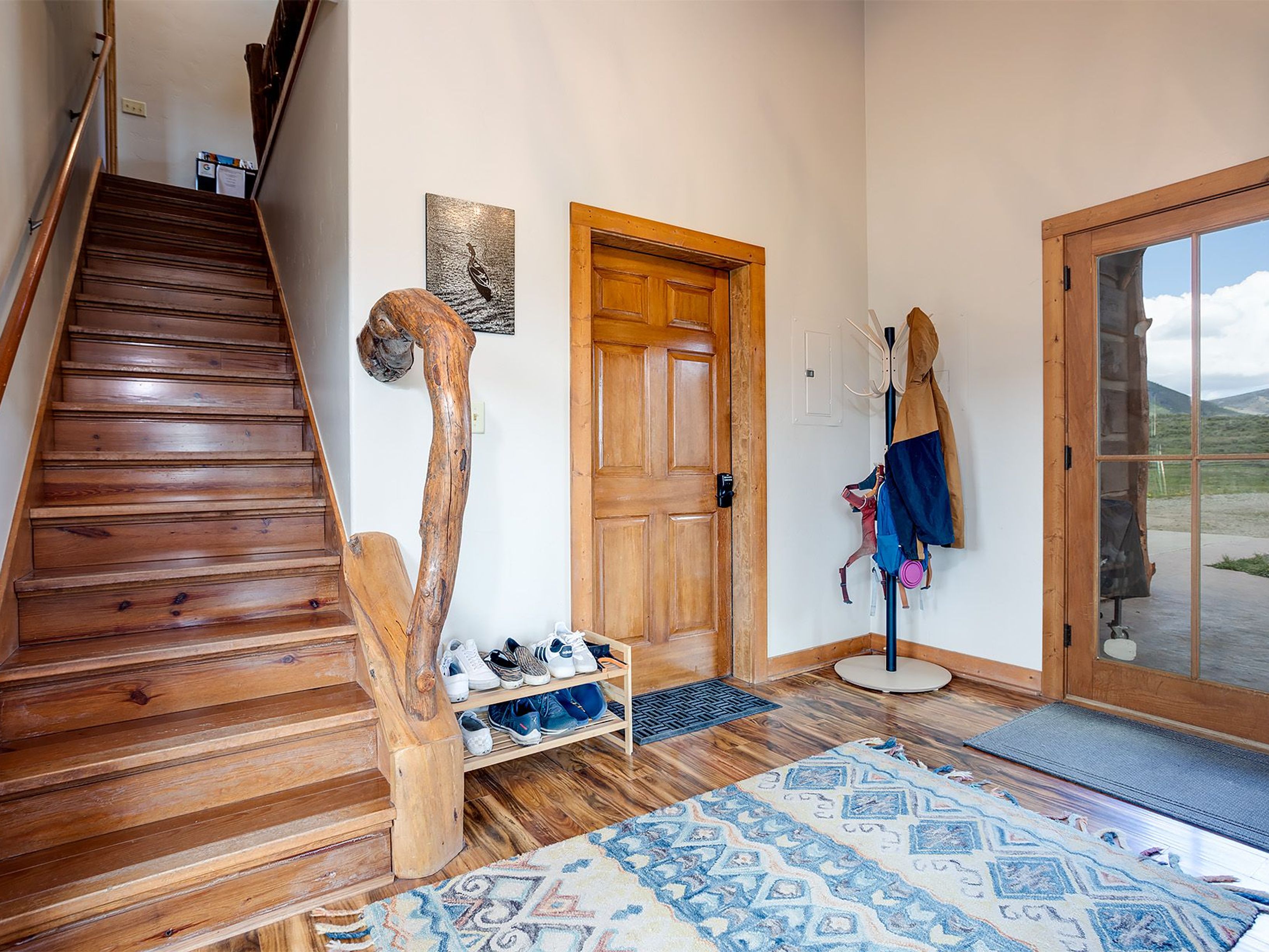 This is an interior shot of a hallway or entryway featuring wooden stairs, doors, and trim. The area is decorated with a unique wooden sculpture-like piece next to a shoe rack, an area rug, and a coat rack. The floor is wood adding warmth and texture to the space.