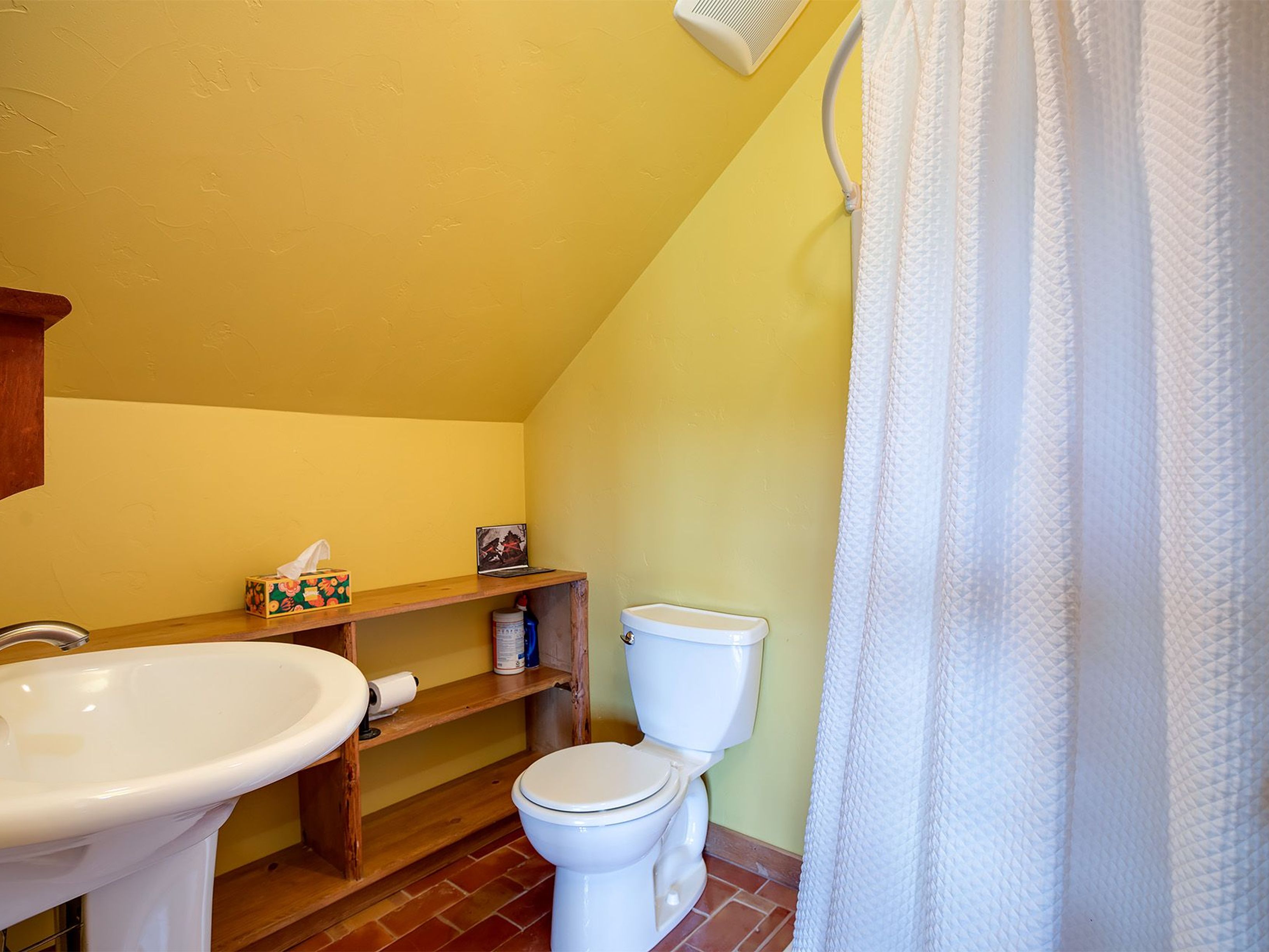 This is a bathroom featuring a white toilet and a pedestal sink with a wooden shelf unit nearby. The walls are painted in yellow, providing a warm tone to the space. A white shower curtain is on the right side that conceals a shower or bathtub.
