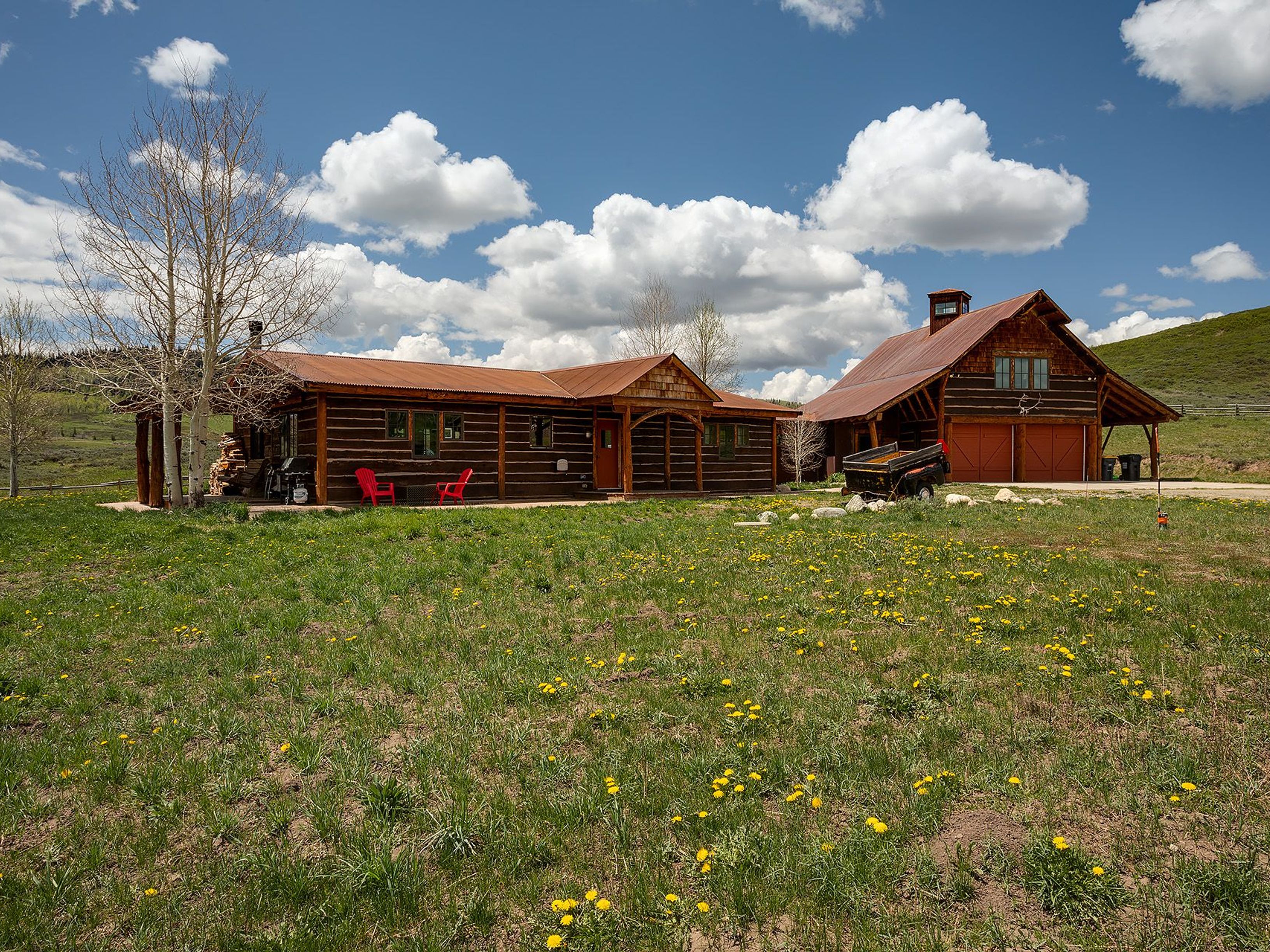 This image showcases the front of a rustic, cabin-style home with a charming barn. The property features a well-maintained lawn with wildflowers, creating a picturesque setting. The log construction, combined with the red-toned metal roof and garage doors, offers a cozy and inviting aesthetic.