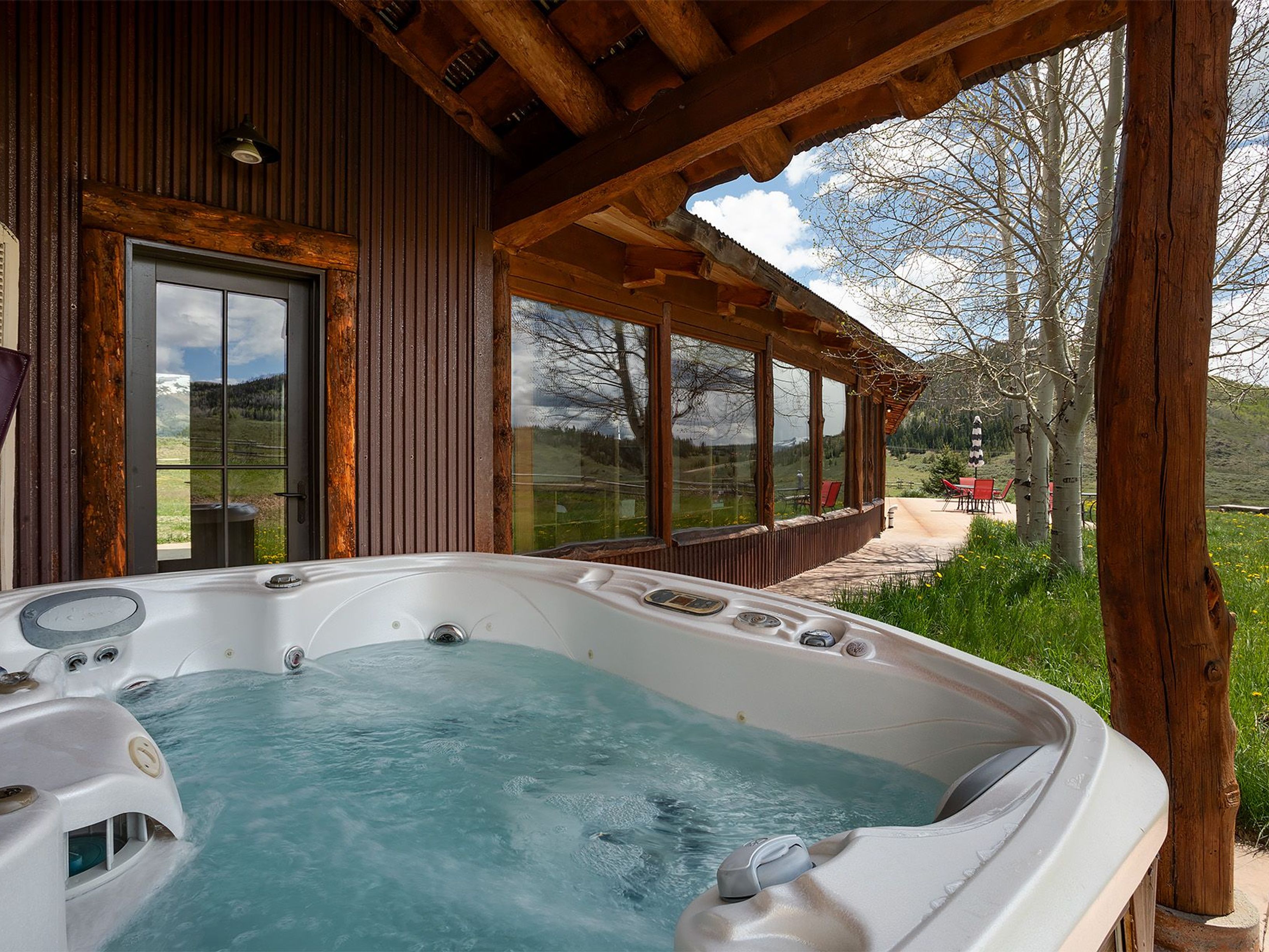 The image showcases a luxurious outdoor hot tub/spa. It's situated on a patio or deck area alongside a property with rustic wooden architecture, providing a sense of privacy and connection with nature. The clean, inviting water and surrounding landscape suggest relaxation and leisure.