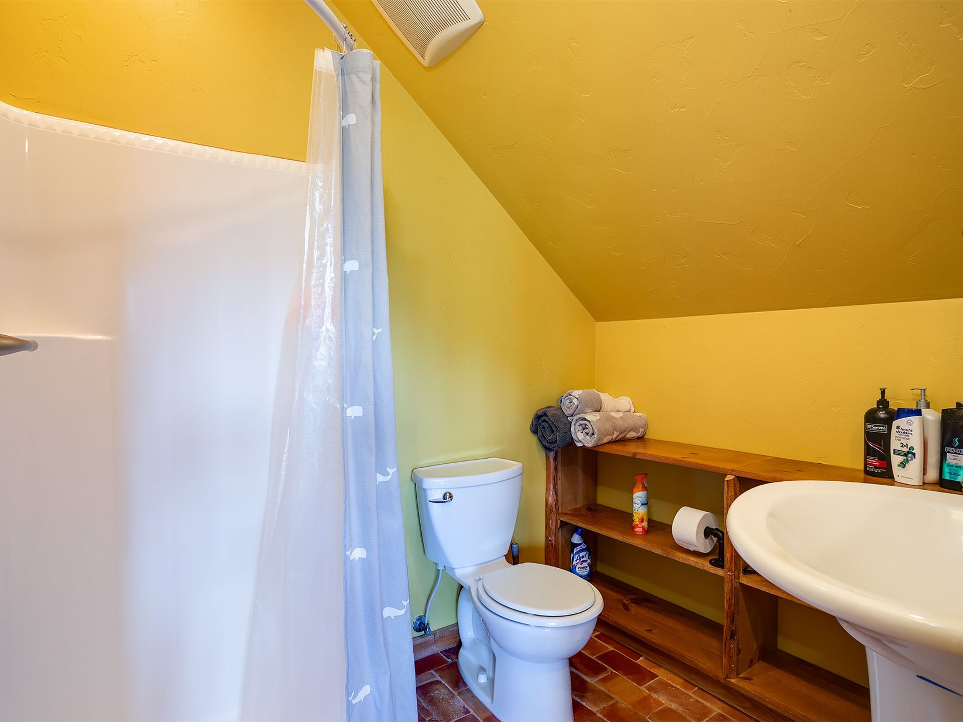 This is a bathroom with ochre walls, showcasing a shower with a curtain, a toilet, a sink, and a wooden shelf. The floor is tiled in reddish-brown, creating a warm and inviting space. The room seems compact but functional with basic sanitary facilities for a home.