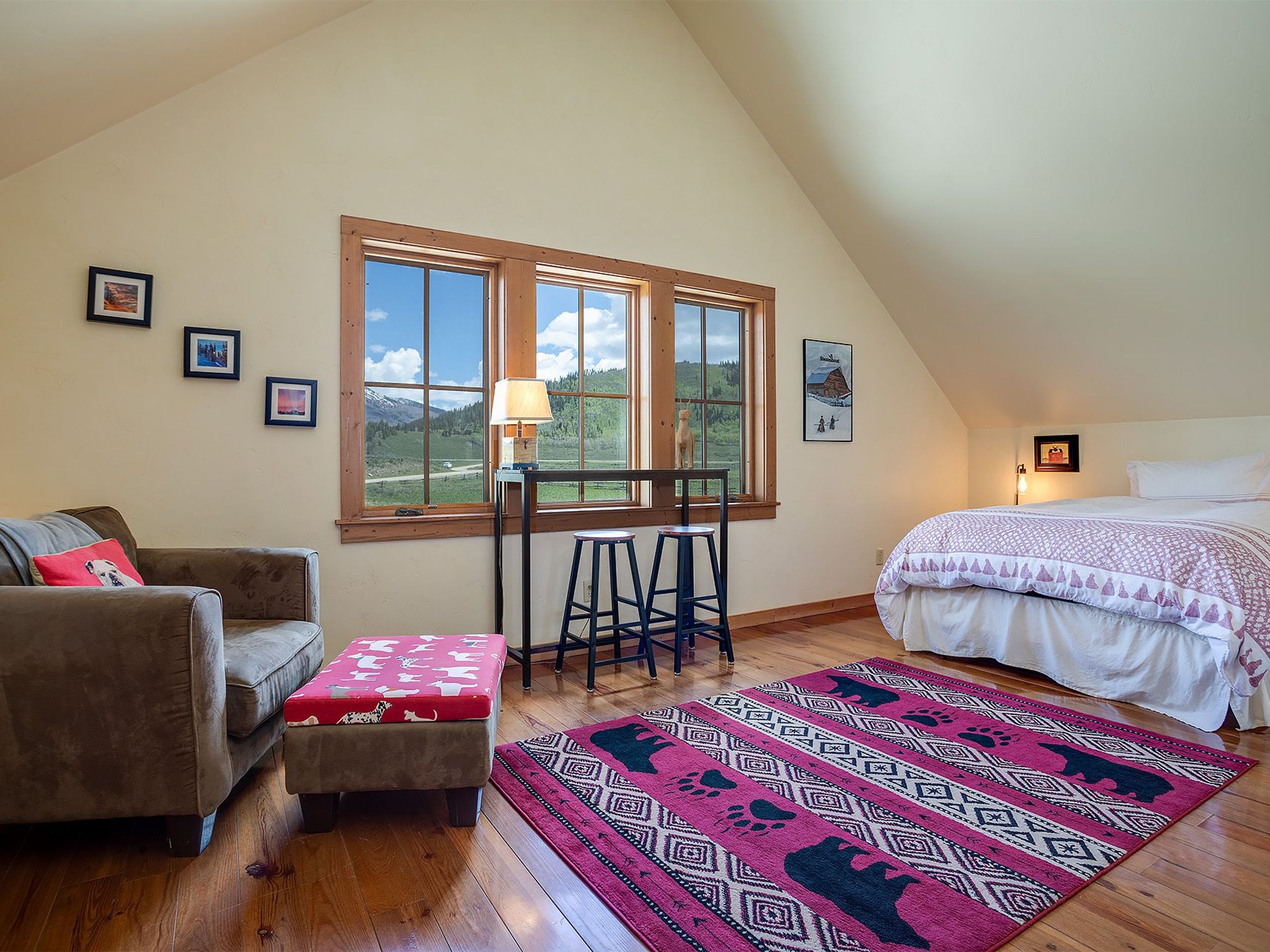 This is a cozy guest bedroom featuring hardwood floors and an area rug with bear motifs. The room is well-lit with natural light coming through multiple windows offering views of the outdoors. Furnishings include a bed, a small seating area with a sofa and ottoman, and a table with stools by the window, making it a comfortable and inviting space.