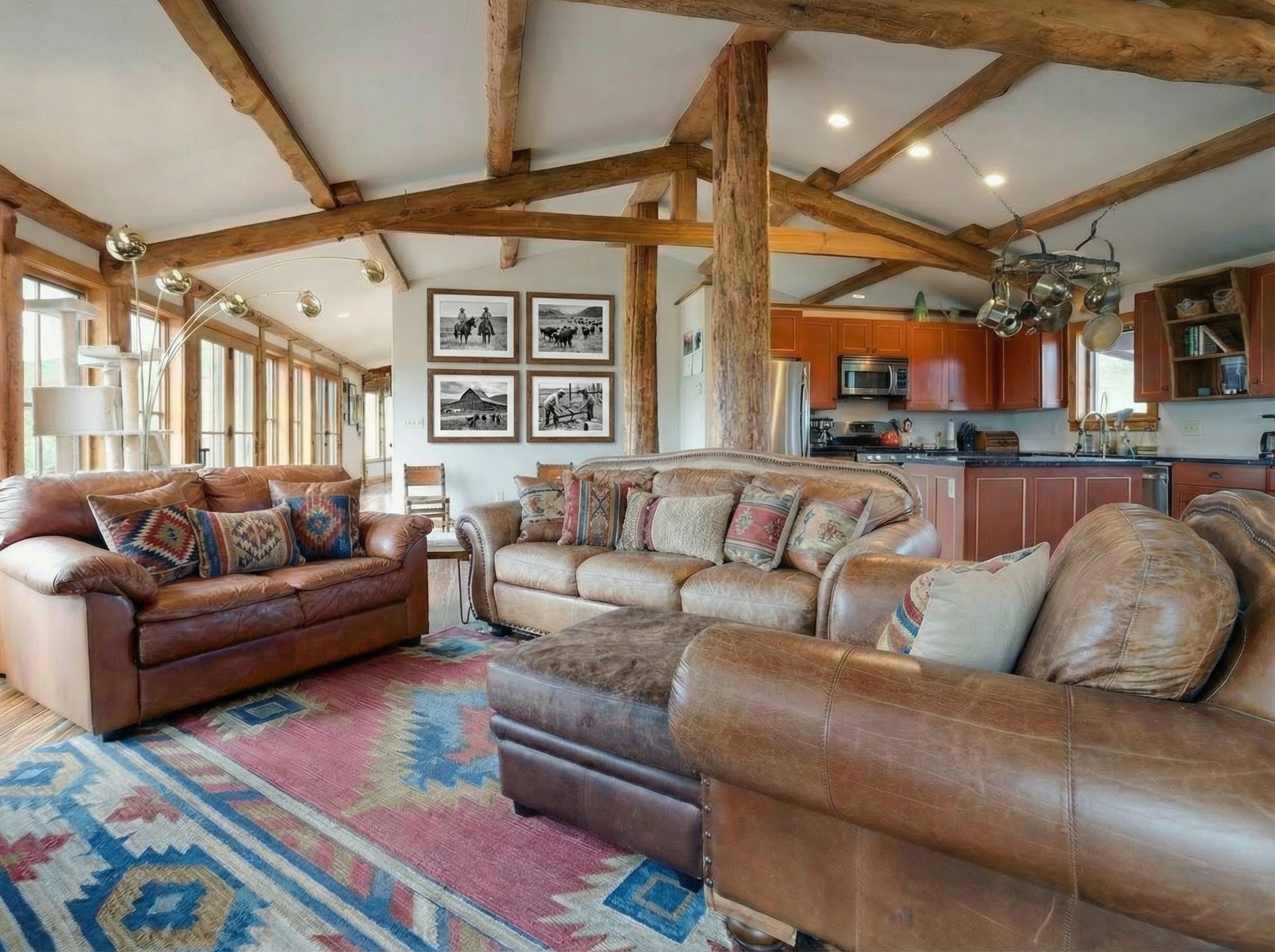 The living room features exposed wood beams and posts, giving a rustic and warm ambiance. Comfortable leather sofas with patterned pillows sit on a colorful area rug. The space opens to the kitchen, creating a cozy yet spacious feel ideal for gatherings.