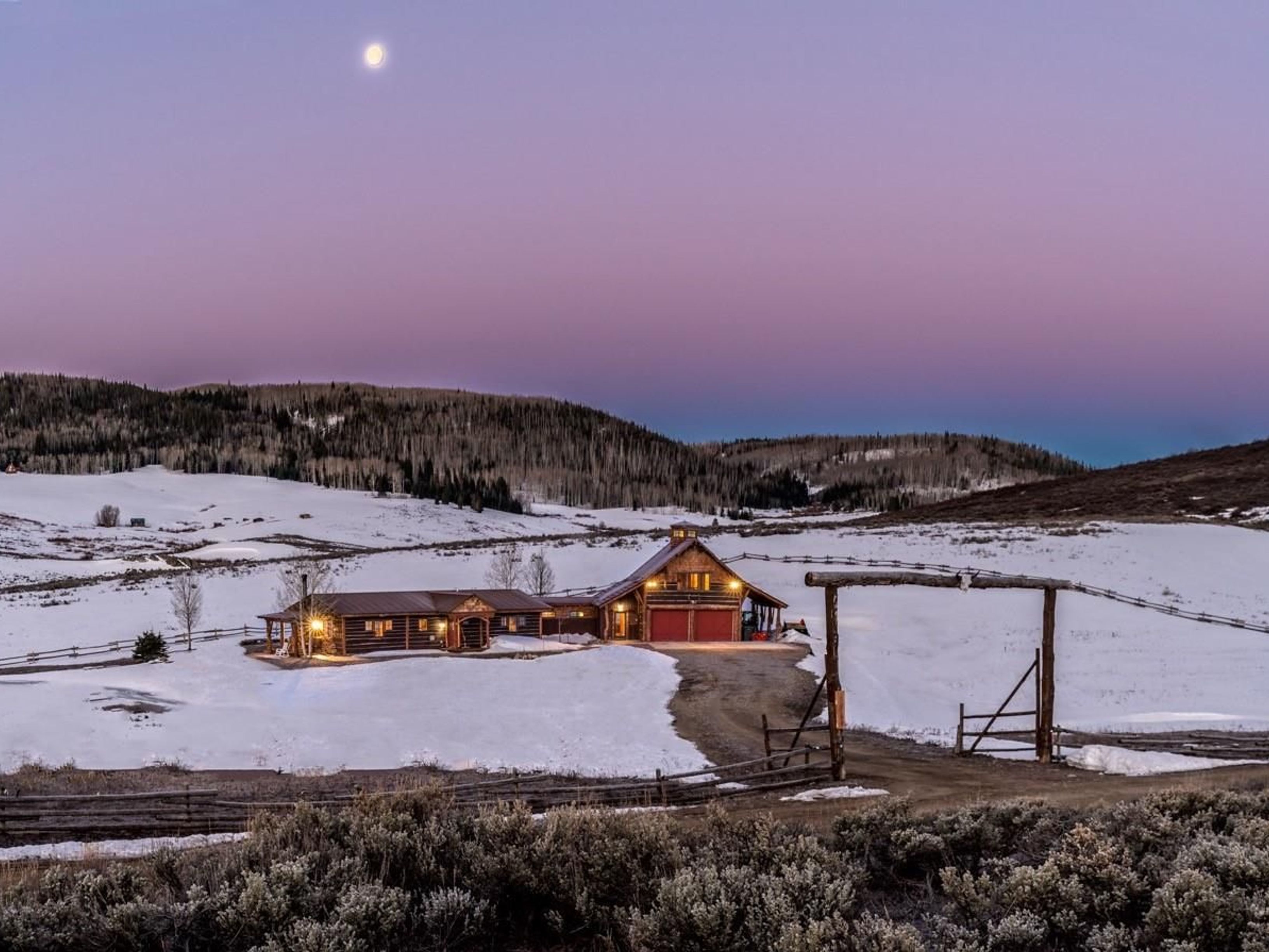 This is an exterior front view of a log cabin home nestled in a snow-covered landscape at dusk. The house features warm interior lighting, red garage doors, and an inviting wooden gate. The property is surrounded by hills and a few trees under a vibrant twilight sky with the moon visible.