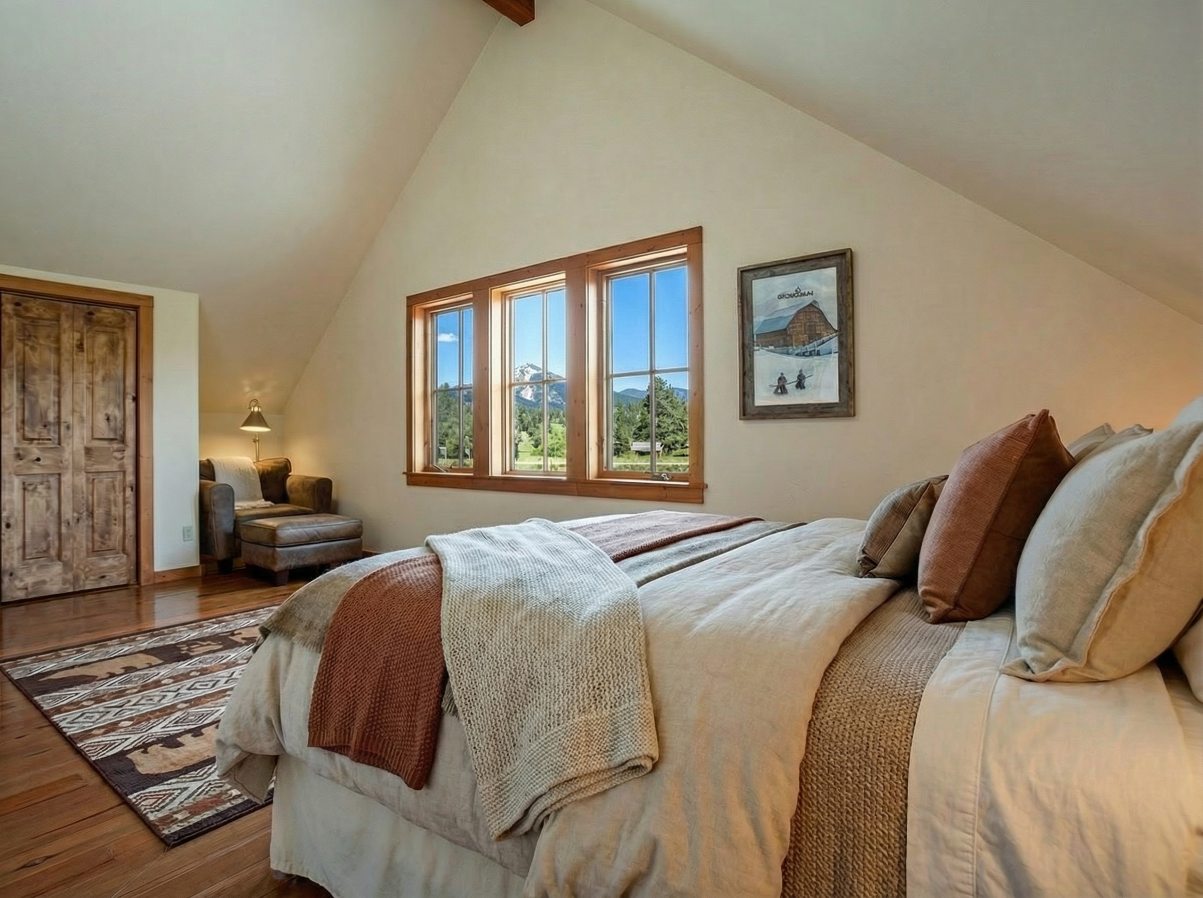This is a cozy primary bedroom in a home with a sloped ceiling. The room is well-lit by three windows that showcase an outdoor view. The room is decorated with neutral tones and features a decorative rug, a brown leather chair, and a framed piece of artwork on the wall.
