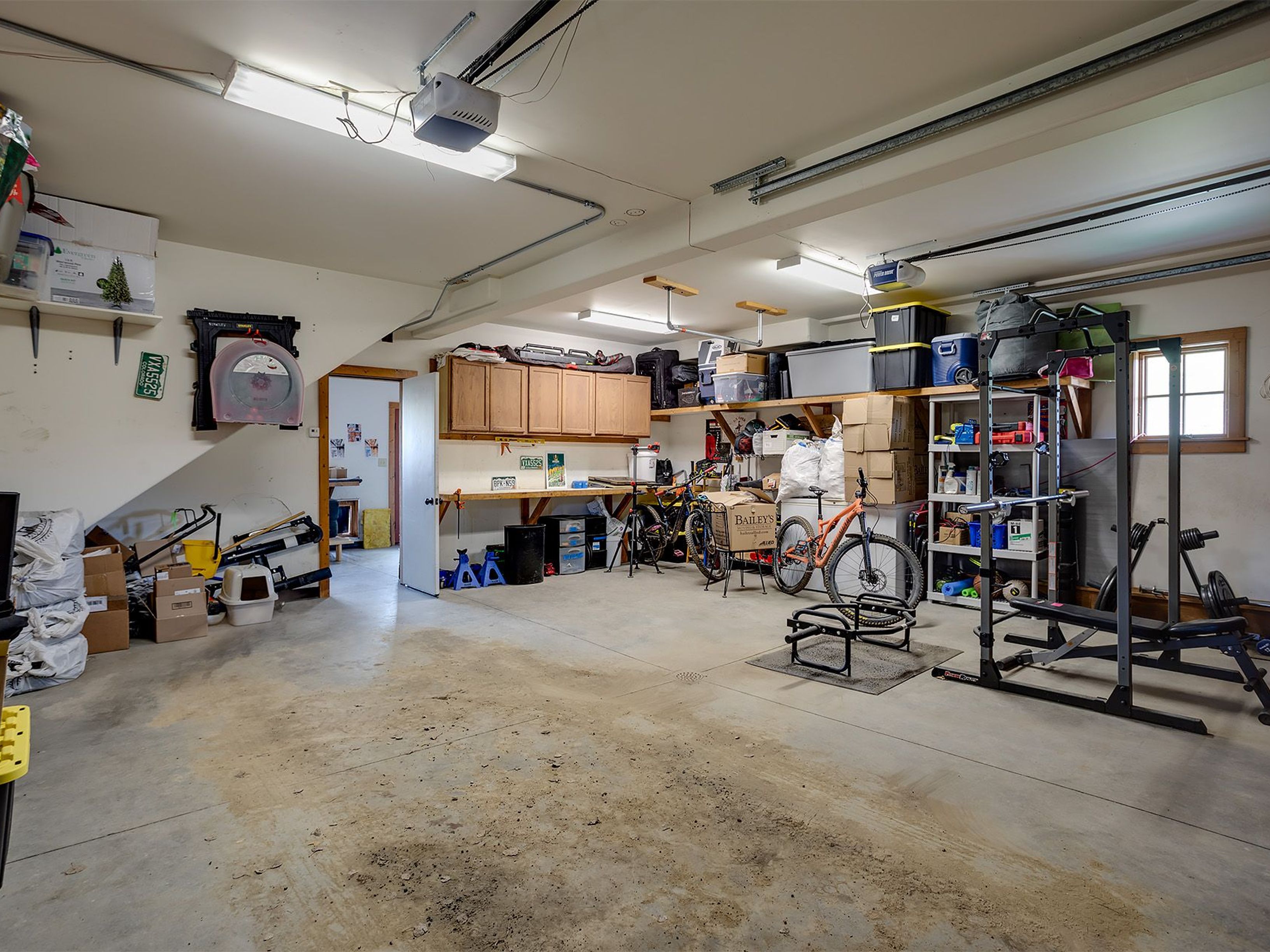 This is a spacious garage with a concrete floor and a variety of storage solutions. Several bikes are visible, along with a full weight set in the corner and cabinets with countertop workspace. The room is well-lit and appears functional, suitable for parking, storage, and hobbies.