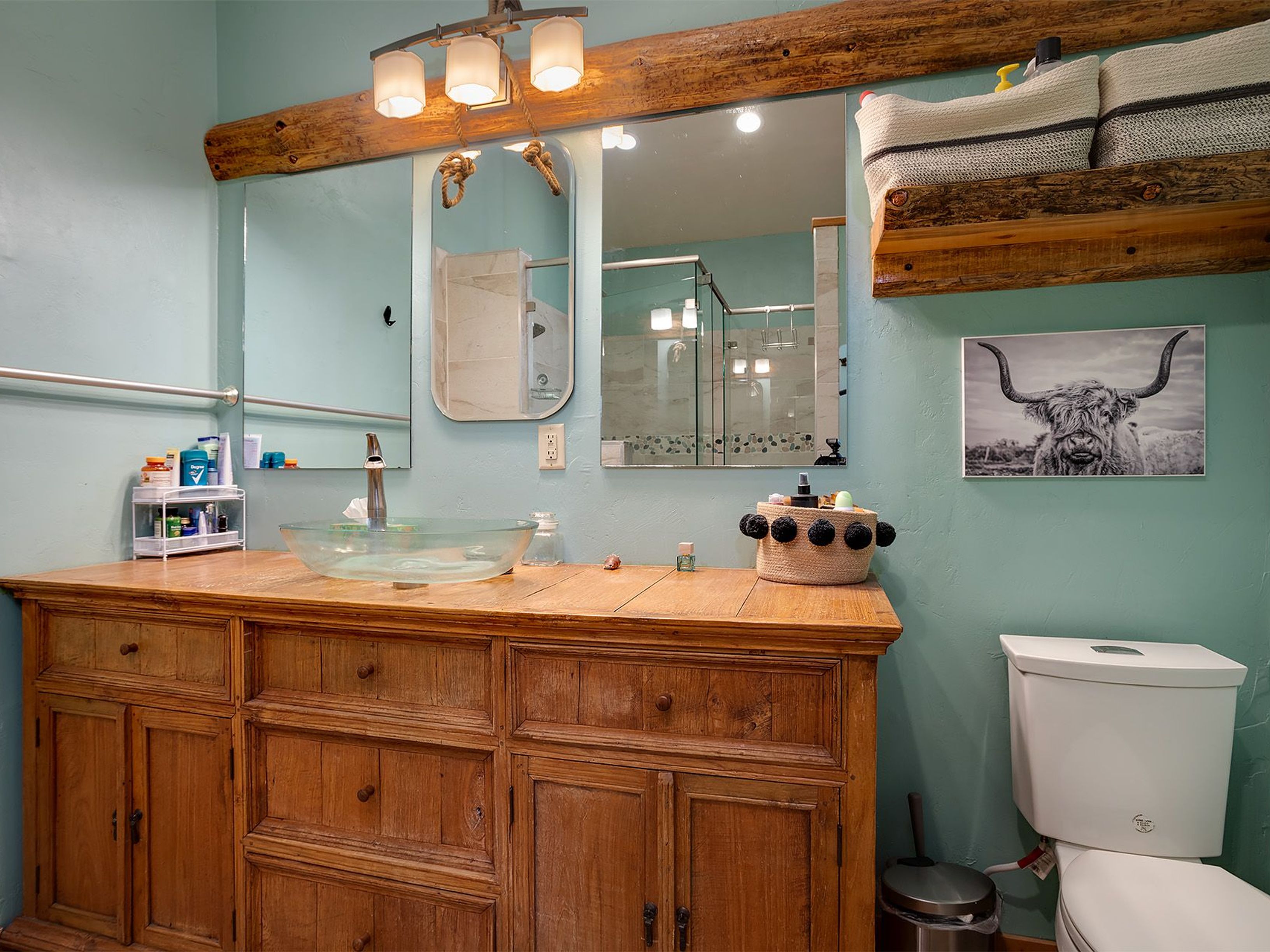 This is a well-lit bathroom featuring a rustic-style wooden vanity with a clear glass vessel sink. The walls are painted in a light aqua color. A decorative mirror hangs above the sink, and a framed print of a Scottish Highland Cow hangs on the wall beside the toilet.