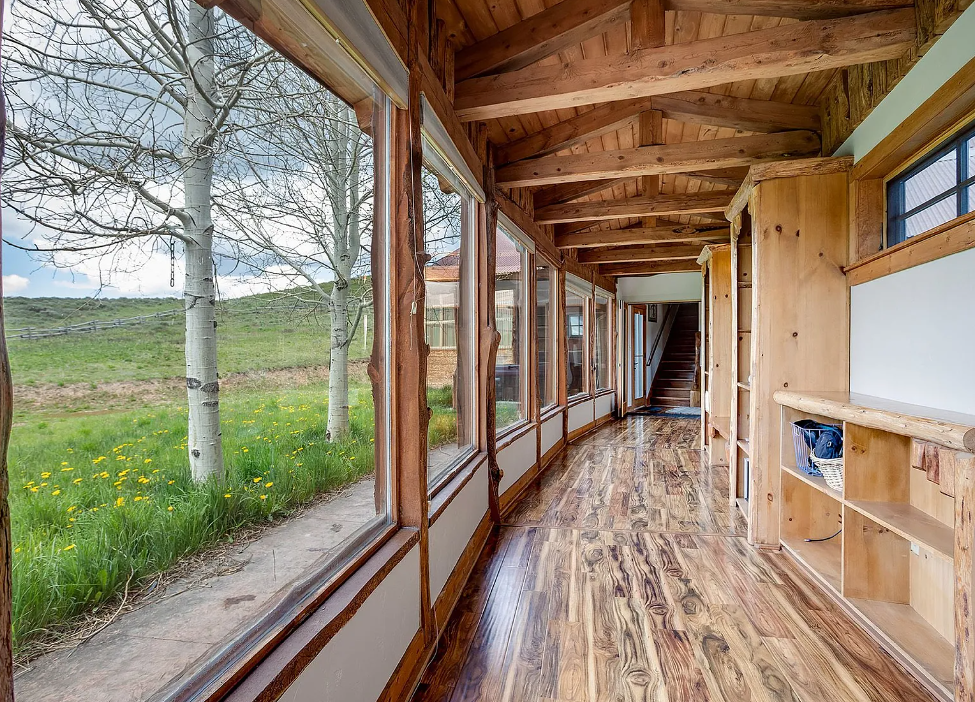 This interior shot showcases a rustic hallway featuring a unique wood floor and timber-framed ceiling. Large windows offer a scenic view of the outdoors, while a built-in wooden shelving unit provides storage. A staircase is visible in the background, suggesting a multi-level home.