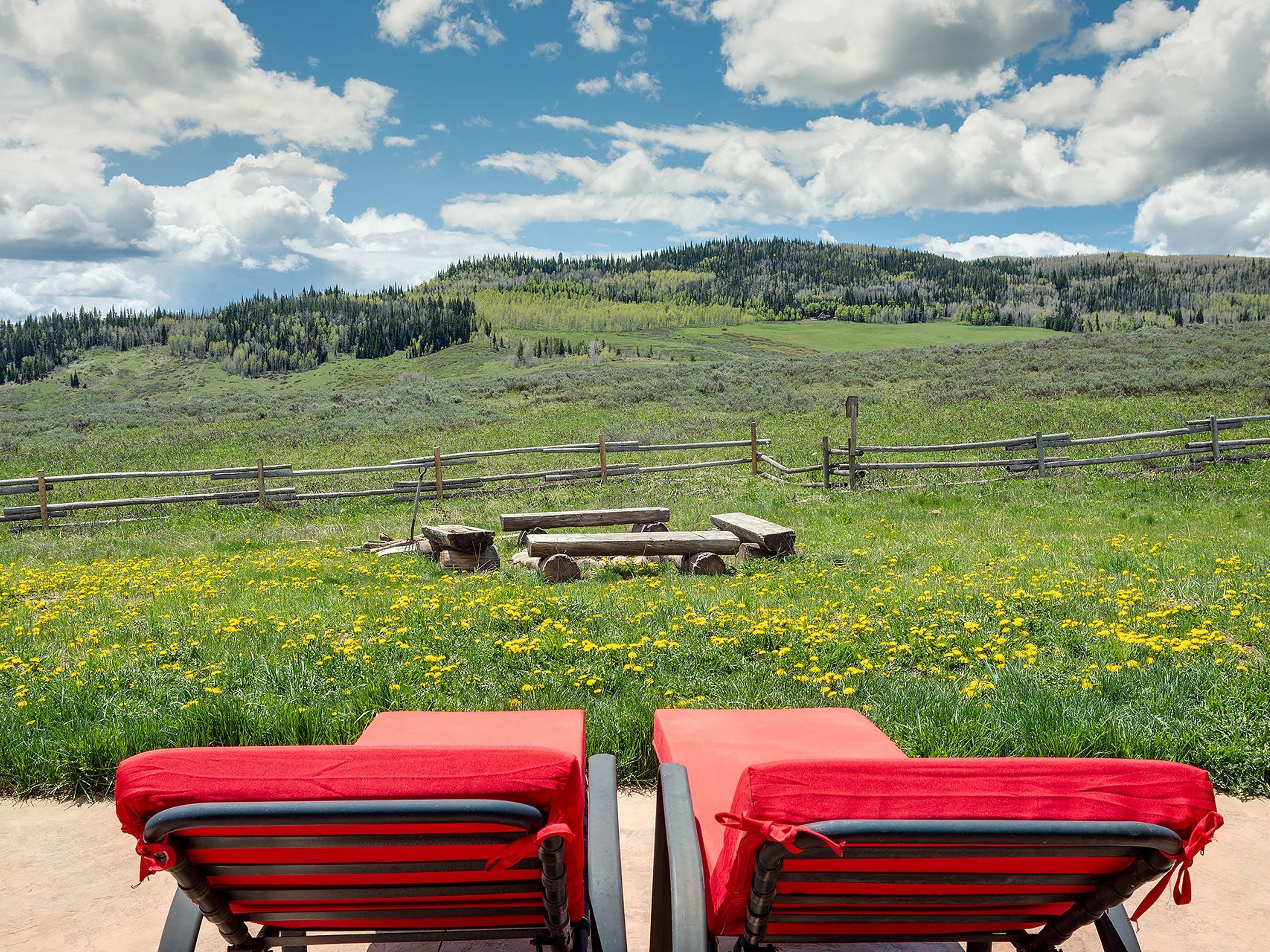 This image showcases an inviting outdoor patio area. Two red-cushioned lounge chairs sit on the patio, facing a picturesque scene with a wooden seating area, a meadow of wildflowers, a rustic wooden fence, and a forested hillside under a bright blue sky with puffy clouds. The scene evokes a sense of tranquility and natural beauty, perfect for relaxation and enjoying the outdoors.