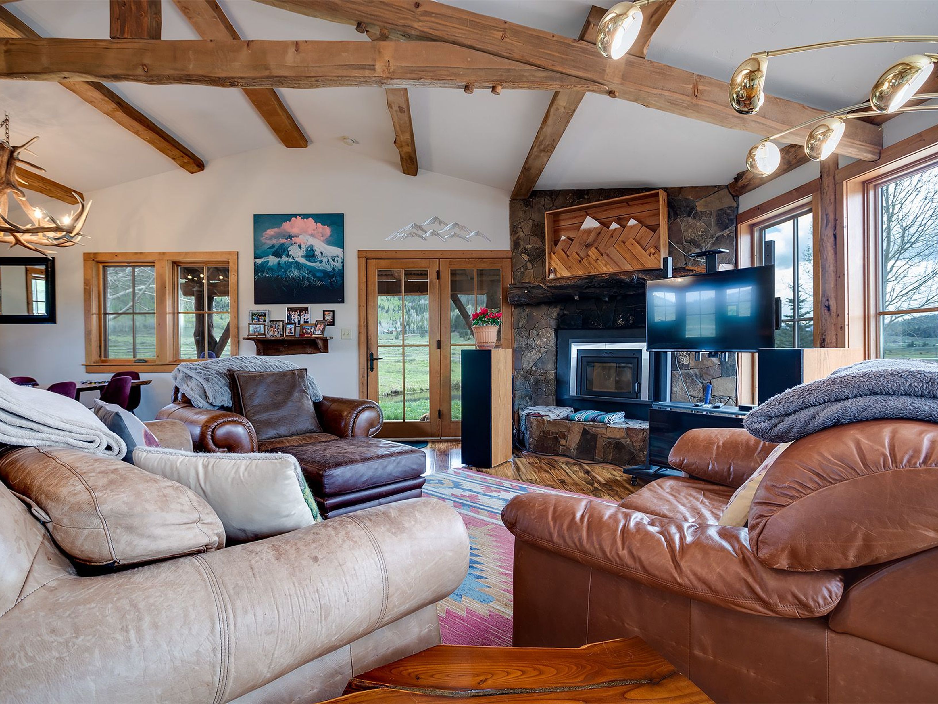This living room features exposed wooden beams, a stone fireplace with a wooden mantel art piece, and leather armchairs. A patterned rug adds warmth to the room, while large windows provide natural light and views of the outdoors. The overall style is rustic and inviting.