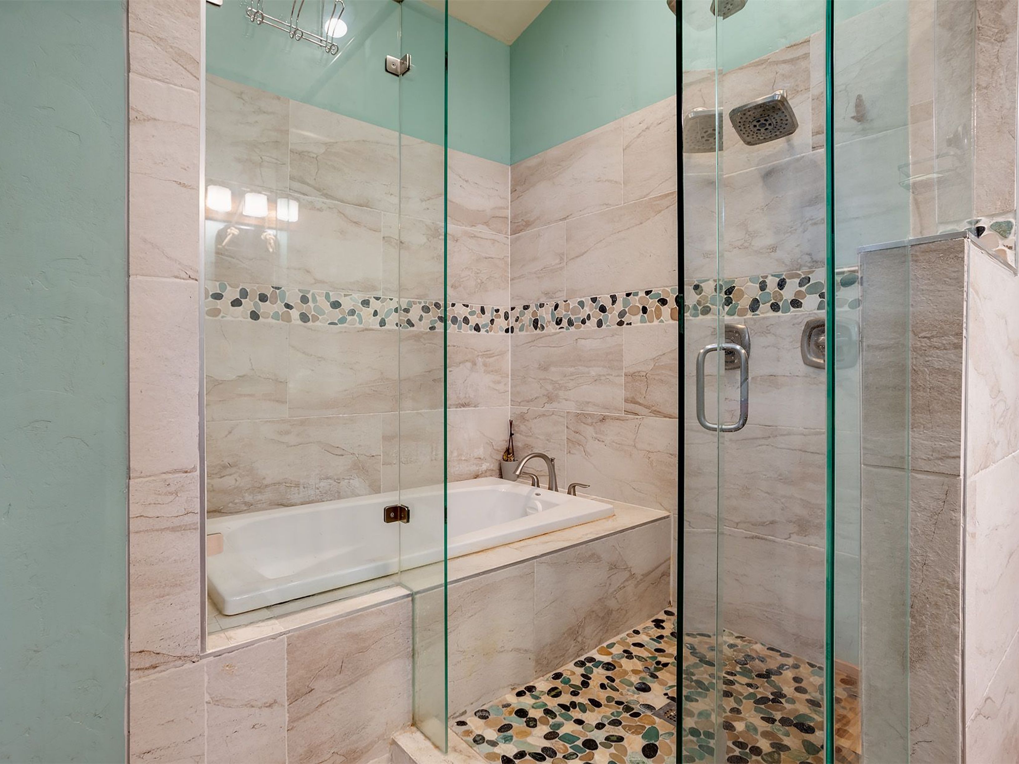 This is a shot of a primary bath, showing the glass enclosed shower and tub area. The walls are a neutral stone tile and the floor of the shower features a unique cobblestone design. The wall above the tile is painted a complementary light green.
