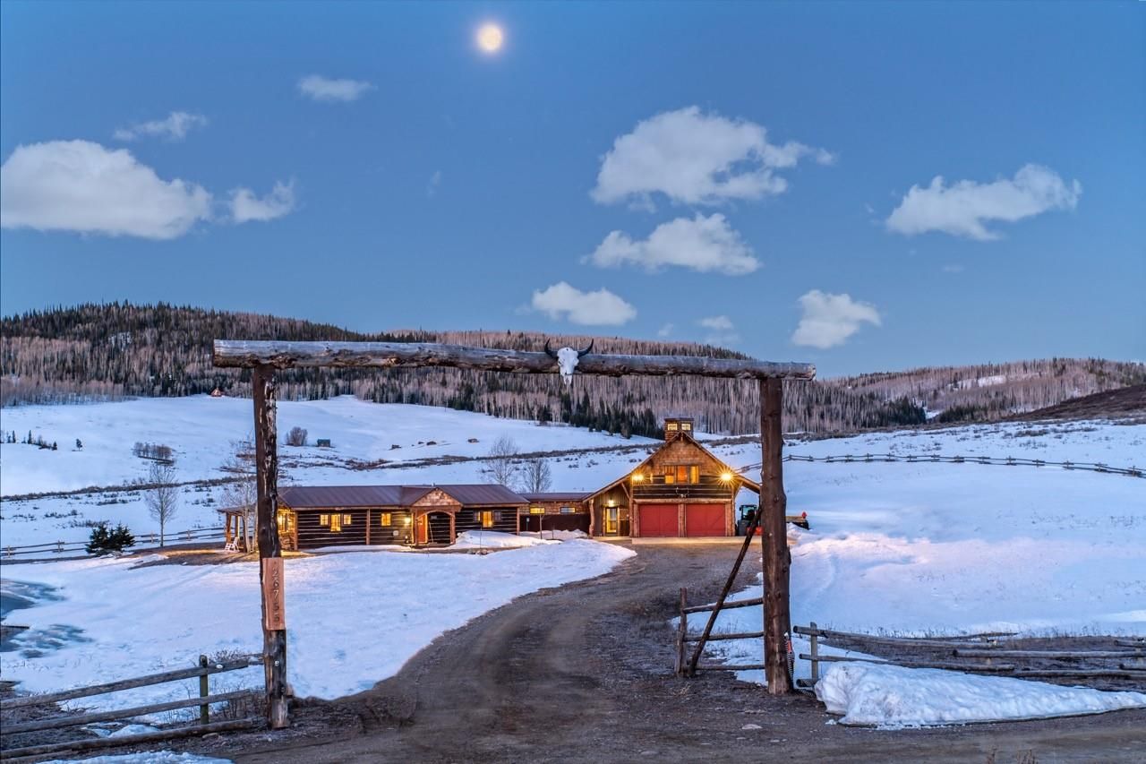 This is an inviting front view of a rustic property in a snowy landscape. It features a log home connected to a barn with red garage doors and a wooden entrance archway adorned with a skull. The scene is picturesque with a full moon and a sky with clouds, suggesting a serene and private setting.