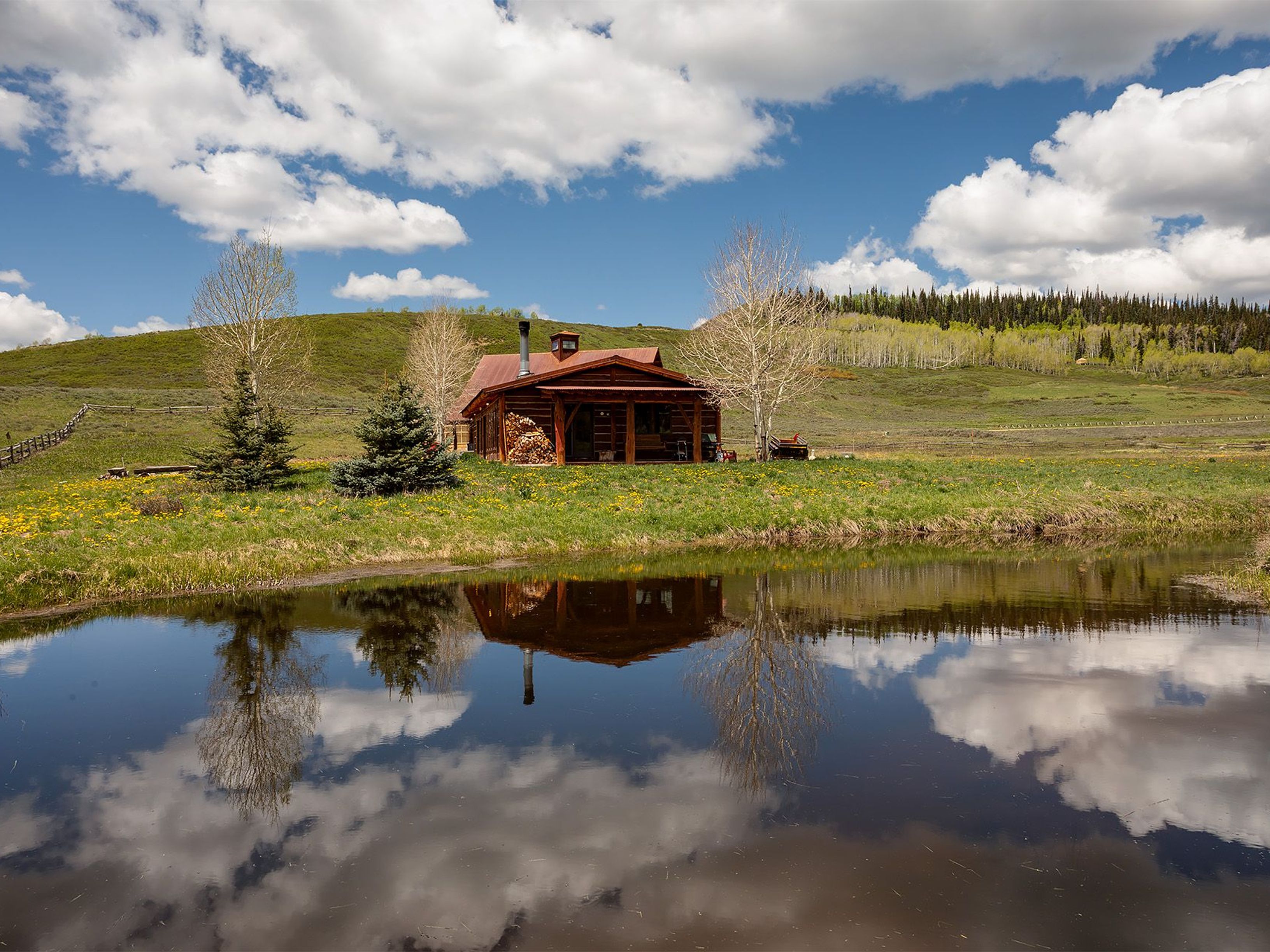 This image showcases the beautiful front exterior of a log cabin home reflecting off a still pond. The cabin features a wooden structure, rustic roof, and a stack of firewood, suggesting a cozy retreat. The landscape includes a green meadow, trees, and a partly cloudy sky.