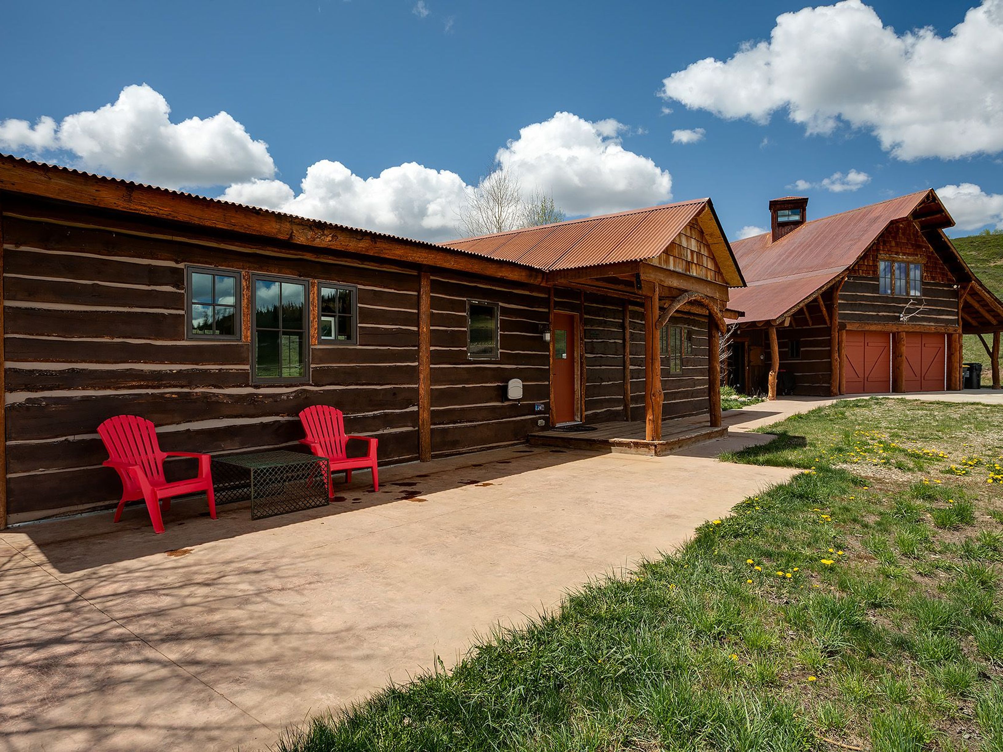 This image captures the front of a charming log cabin home. The property features a rusted metal roof, a spacious concrete patio area with red Adirondack chairs, and well-maintained landscaping with some yellow flowers in the lawn. The house has a rustic aesthetic with exposed log construction and a covered porch.