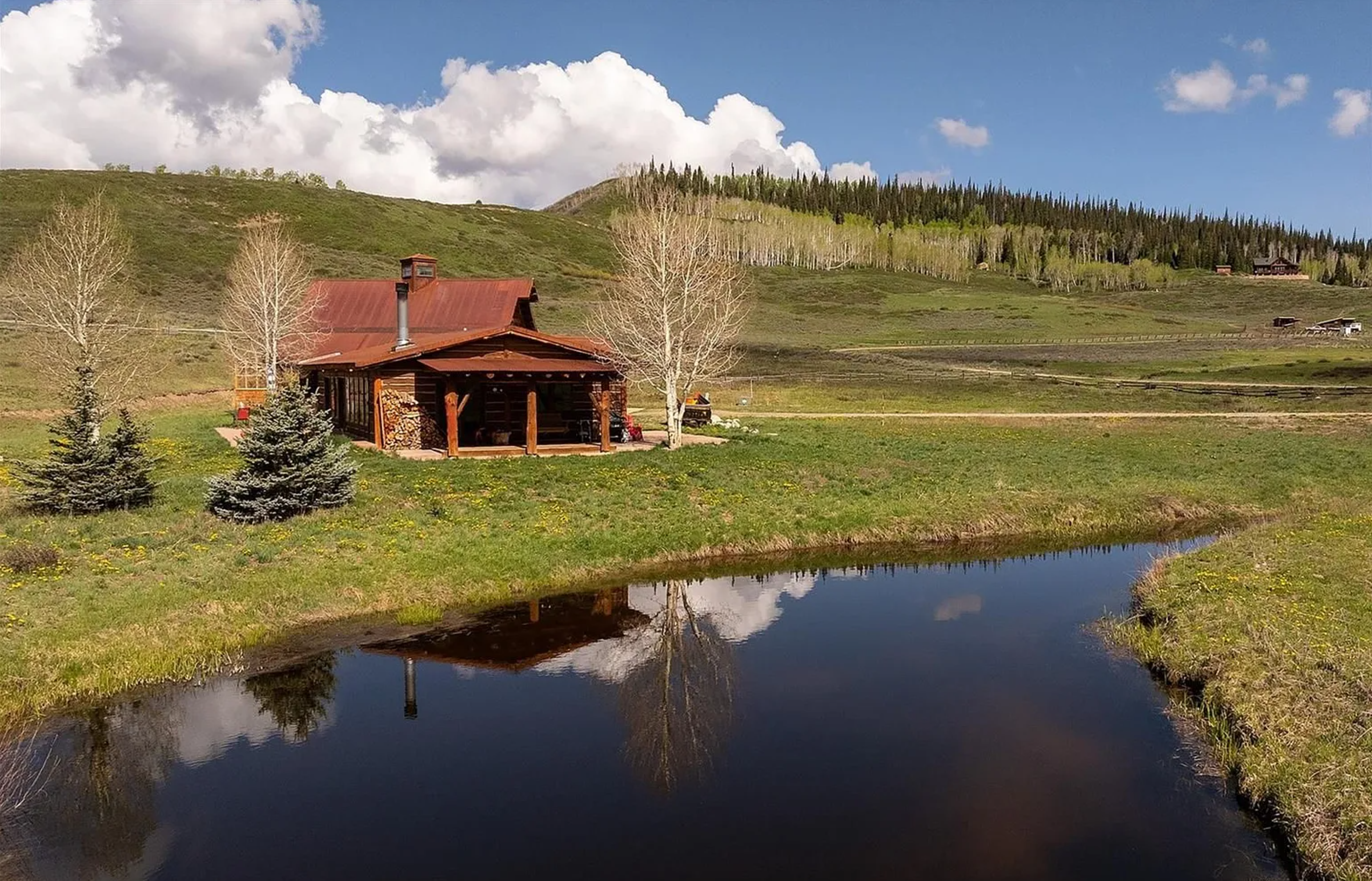 This stunning aerial image showcases a rustic cabin nestled in a picturesque landscape. The cabin features a rust-colored roof, stone accents, and a charming covered porch. A tranquil pond reflects the cabin and surrounding trees, adding to the serene and inviting atmosphere. The property boasts expansive green space and rolling hills with scattered trees, creating a beautiful rural retreat.