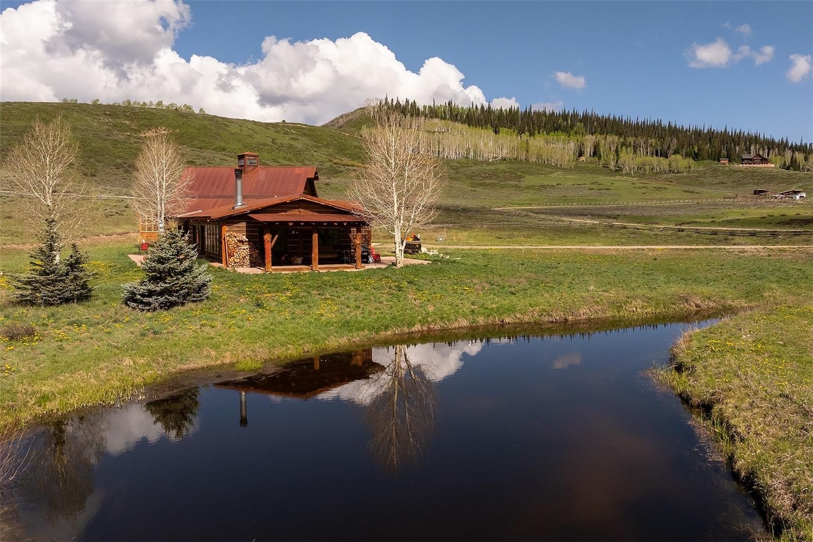 This image showcases the charming front view of a rustic cabin home situated beside a pond. The cabin features a distinctive reddish-brown roof and what appears to be stone and wood siding. The landscape includes green rolling hills, trees, and a clear sky with puffy clouds, emphasizing the home's secluded, nature-filled setting.