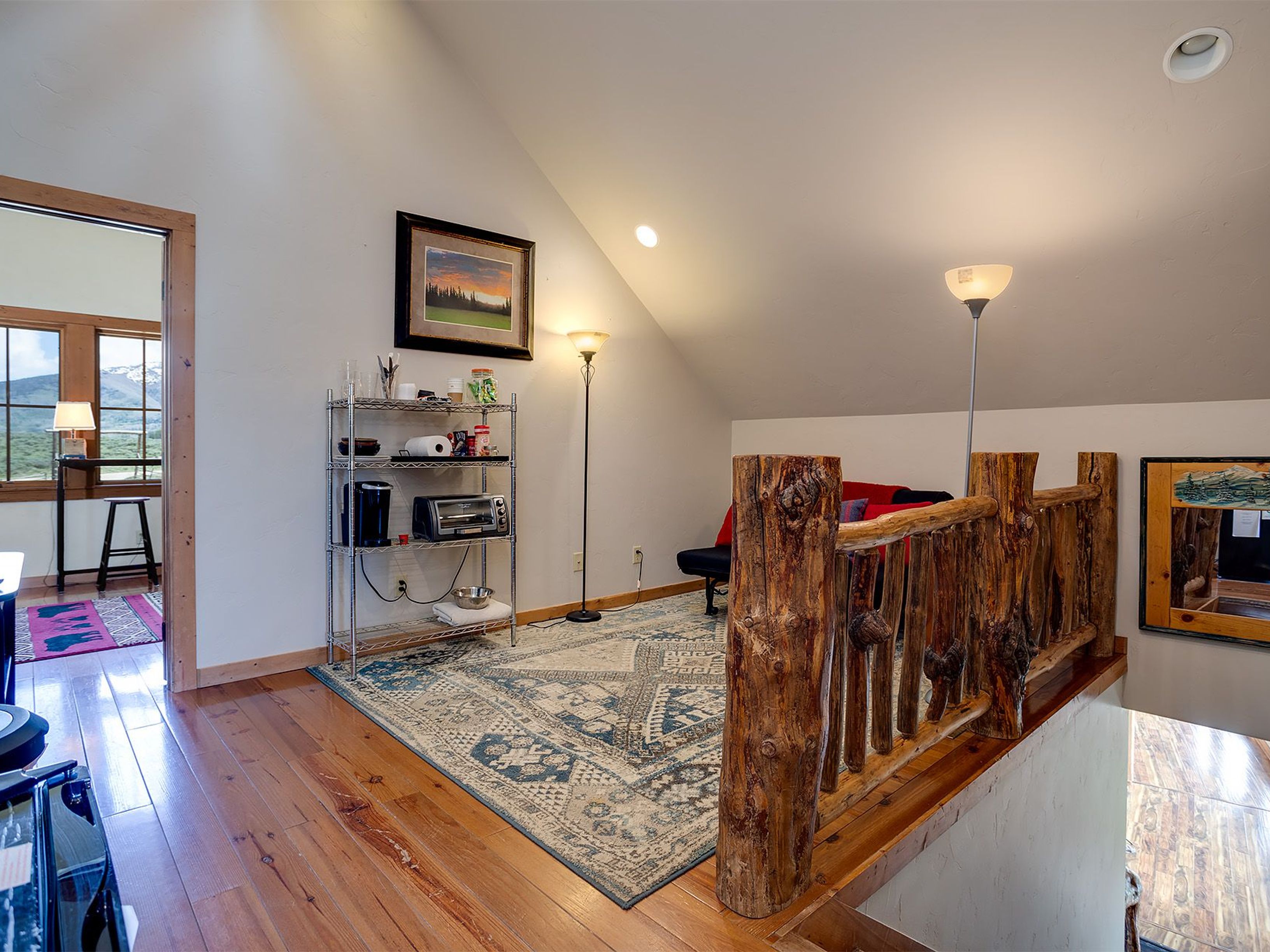 This image showcases a cozy attic space, likely converted into a living area or loft. It features hardwood floors, a rustic wooden railing, and a metal shelving unit. The space is illuminated by floor lamps, creating a warm and inviting atmosphere, which would appeal to potential buyers looking for unique character.