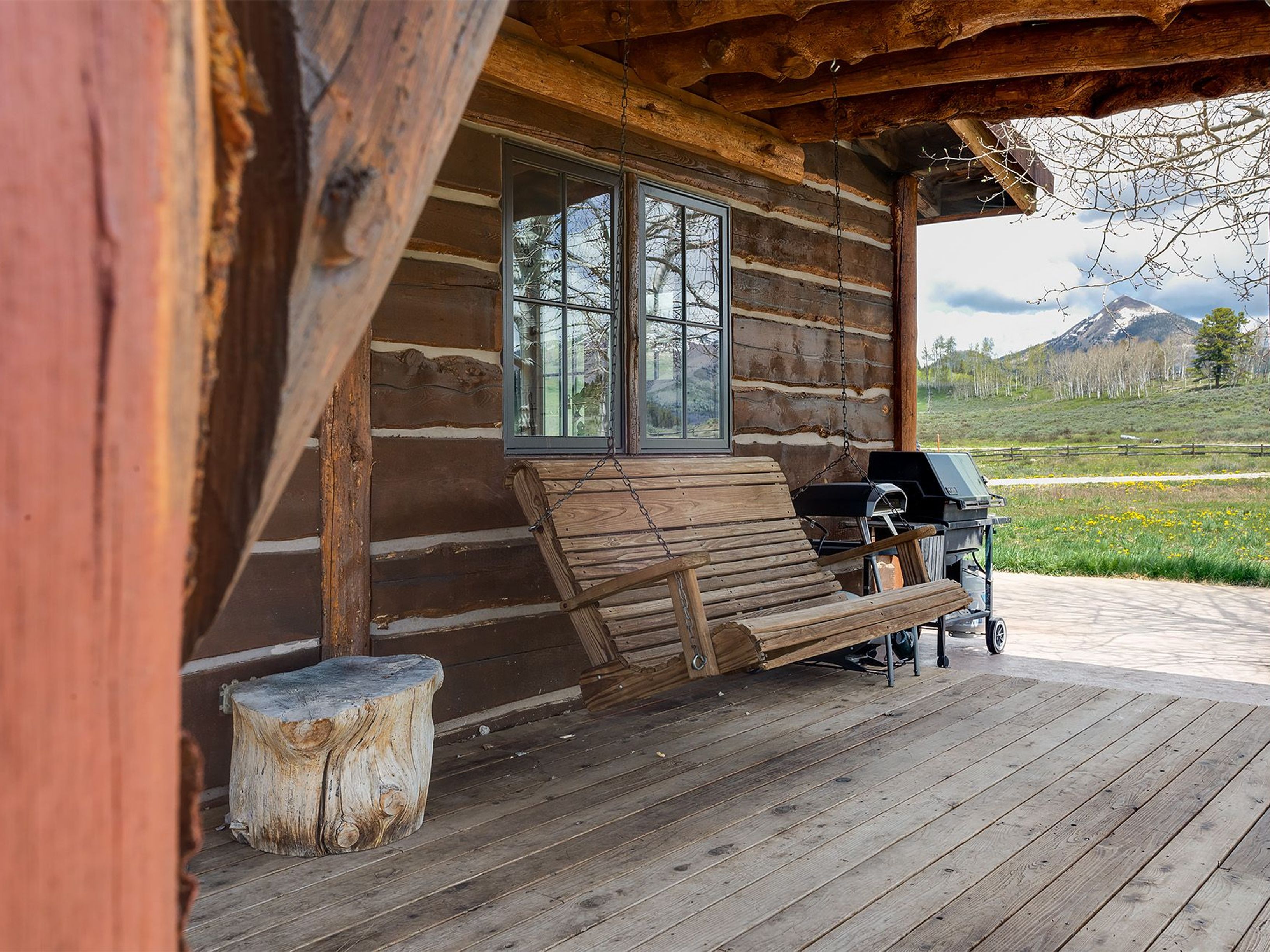 This image showcases a charming patio or deck area attached to a log cabin. A rustic wooden swing hangs invitingly, with a grill positioned nearby, suggesting a perfect space for outdoor relaxation and entertaining. The wooden flooring of the deck and the natural log construction of the cabin contribute to a cozy, rural aesthetic.