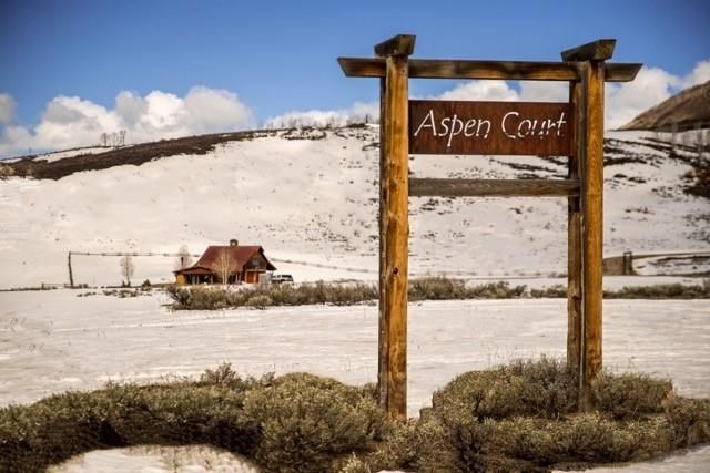 This image showcases a winter scene with an 'Aspen Court' signpost in the foreground, set against a backdrop of snow-covered hills and a clear blue sky. In the middle ground, a cozy-looking house with a reddish-brown roof nestles amidst the snowy terrain, suggesting a secluded and peaceful location. The photograph highlights the property's entrance and its integration with the natural, snowy environment, evoking a sense of rustic charm and winter tranquility.