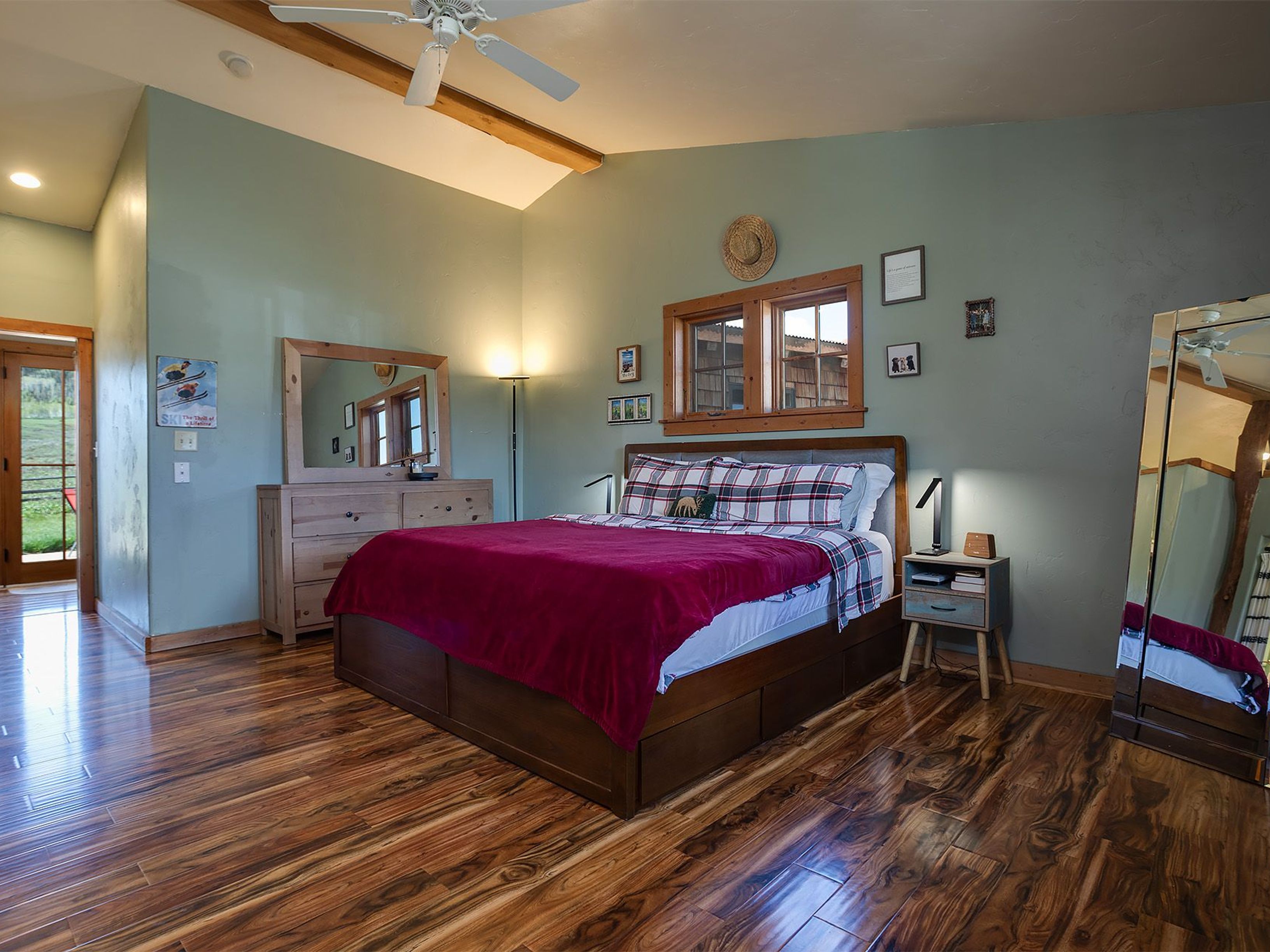 This is a cozy primary bedroom with hardwood flooring and a large bed. The walls are painted in a soft green color. The room features a wooden dresser with a mirror, and there are windows with natural light streaming in, enhancing the warm and inviting atmosphere of the space.