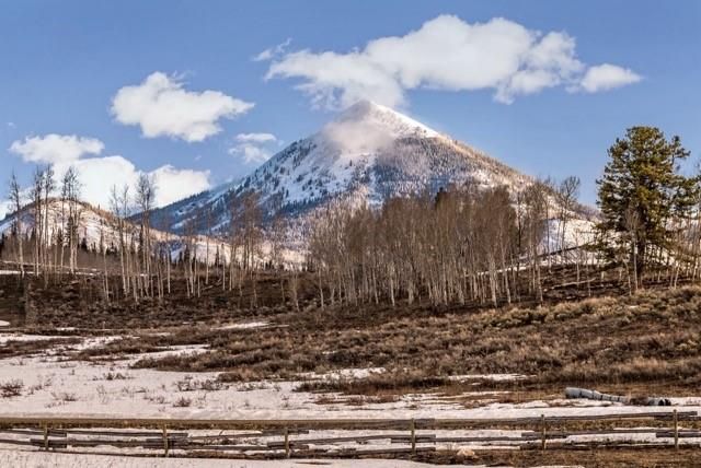 The image showcases a scenic mountain landscape, presumably featuring the front view of a property situated amidst nature. The snow-capped mountain dominates the background, while a mix of trees and open terrain with patches of snow are visible in the foreground. A rustic wooden fence adds a touch of rural charm to the overall view, hinting at a secluded or countryside property.