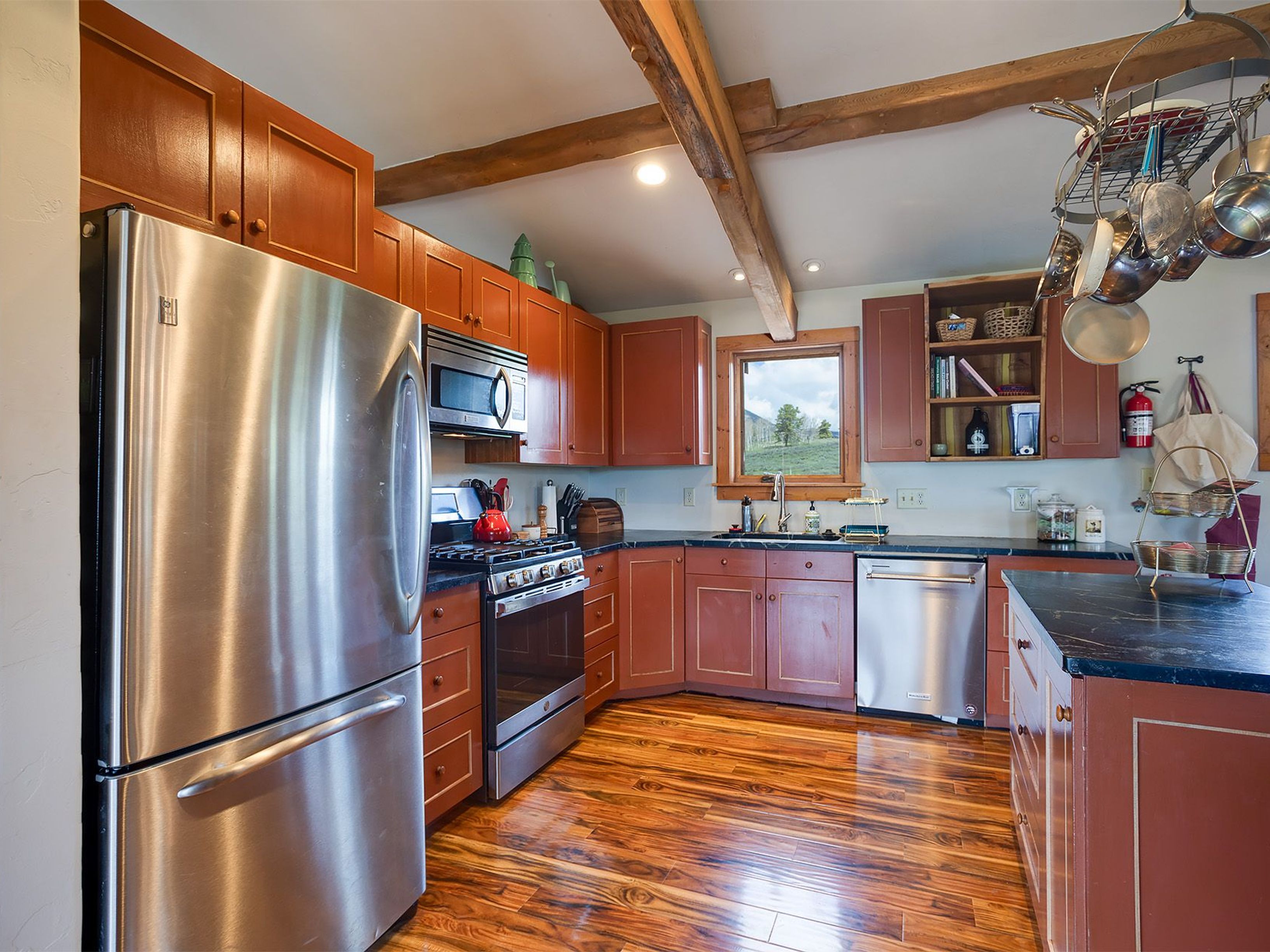 This is a view of a rustic kitchen with wooden cabinets, a stainless steel refrigerator, and wood flooring. Exposed wood beams run across the ceiling, adding to the rustic charm. Overall, the space exudes a warm and inviting atmosphere.