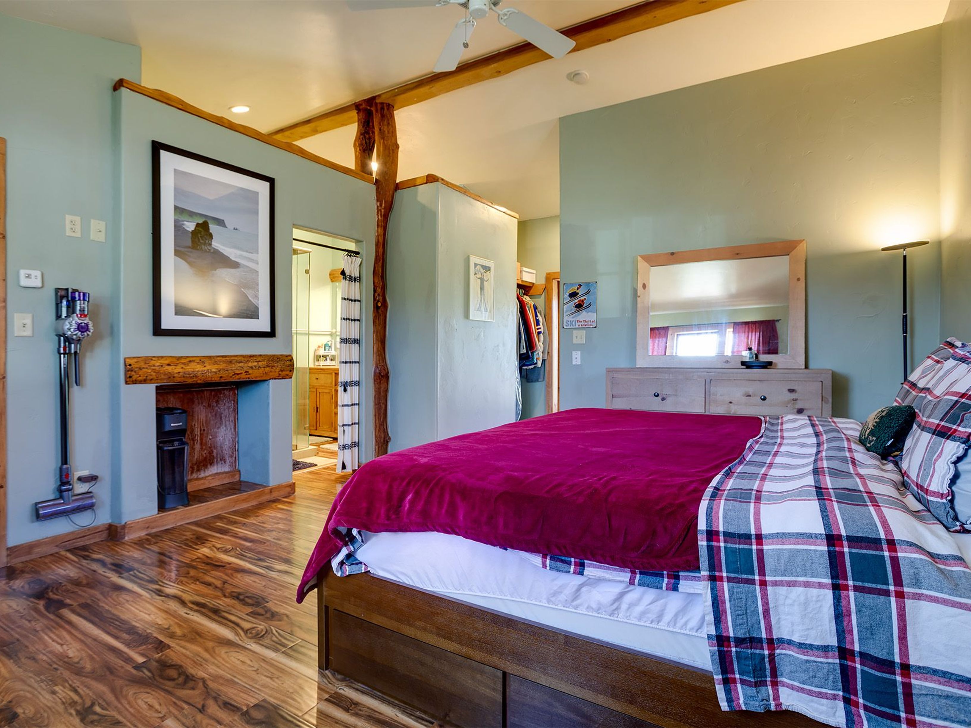 This is a cozy primary bedroom featuring a unique architectural style with exposed wooden beams and a decorative tree trunk support. The room is painted in a calming light green, and the bed is covered with a plush pink blanket and plaid comforter. The furnishings include a wooden dresser with a mirror and a decorative fireplace, creating a warm and inviting space.