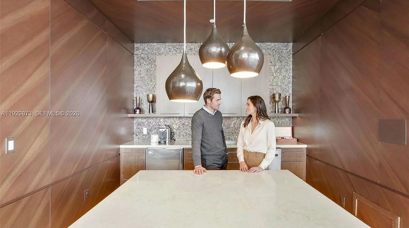 This interior shot showcases a modern kitchen area. The space features a large island with a white countertop, surrounded by warm wood-paneled walls. Three pendant lights hang above the island, illuminating the area where two individuals are standing.