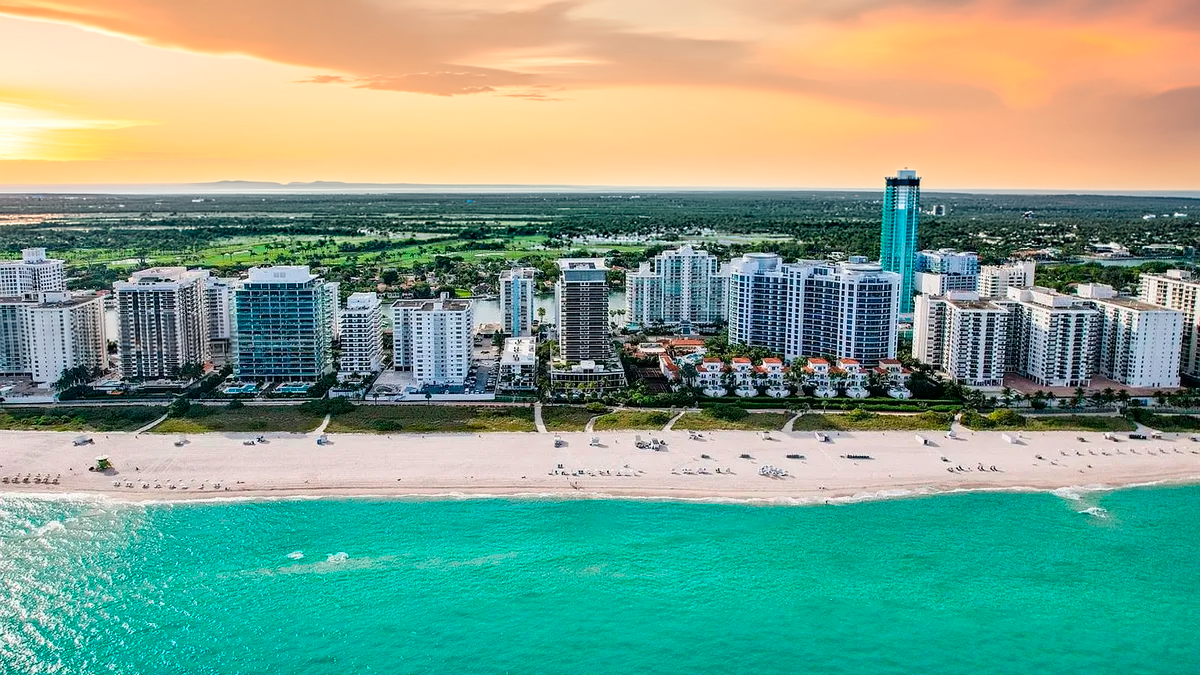 Aerial view of a coastal city featuring numerous high-rise buildings along a sandy beach and turquoise water. The image captures a vibrant sunset with orange and yellow hues in the sky. This view highlights the prime location and stunning waterfront setting of the properties.