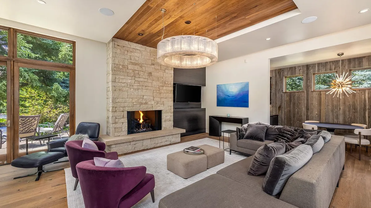 This sophisticated, contemporary living room features a striking floor-to-ceiling stone fireplace as a central focal point, complemented by a warm wood-plank ceiling. The space is furnished with a plush gray sectional, two elegant purple velvet armchairs, and a large circular crystal chandelier. Large glass doors provide seamless indoor-outdoor flow to the patio, enhancing the home's bright and modern aesthetic.